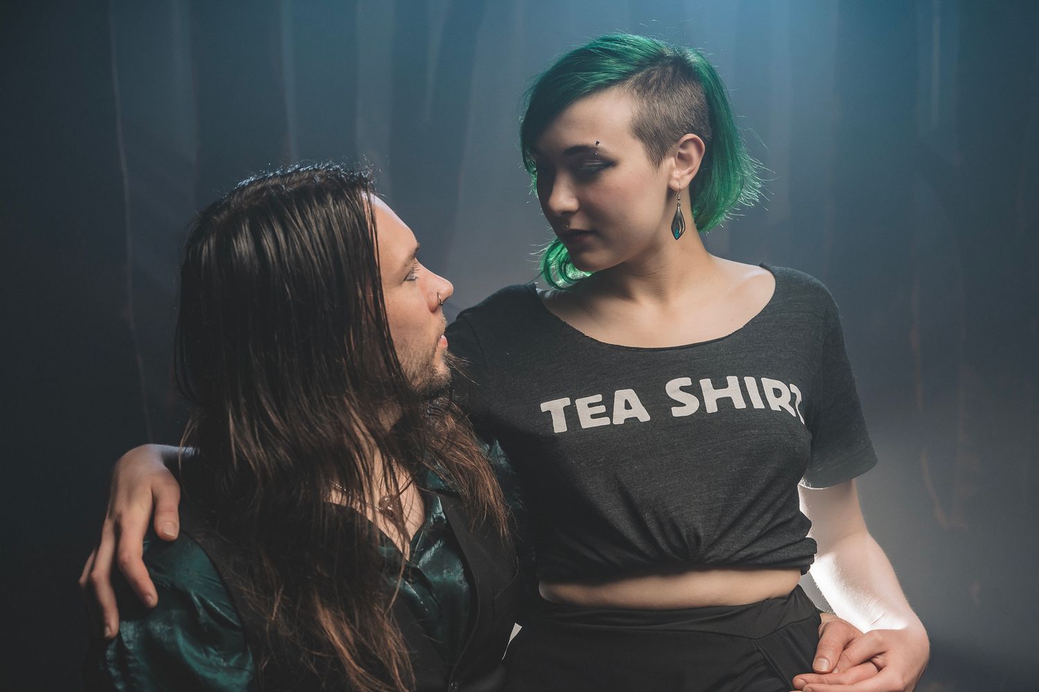 Two women with colorful hair embrace and smile at each other in a dark, moody setting, one wearing a Tea Shirt tee.