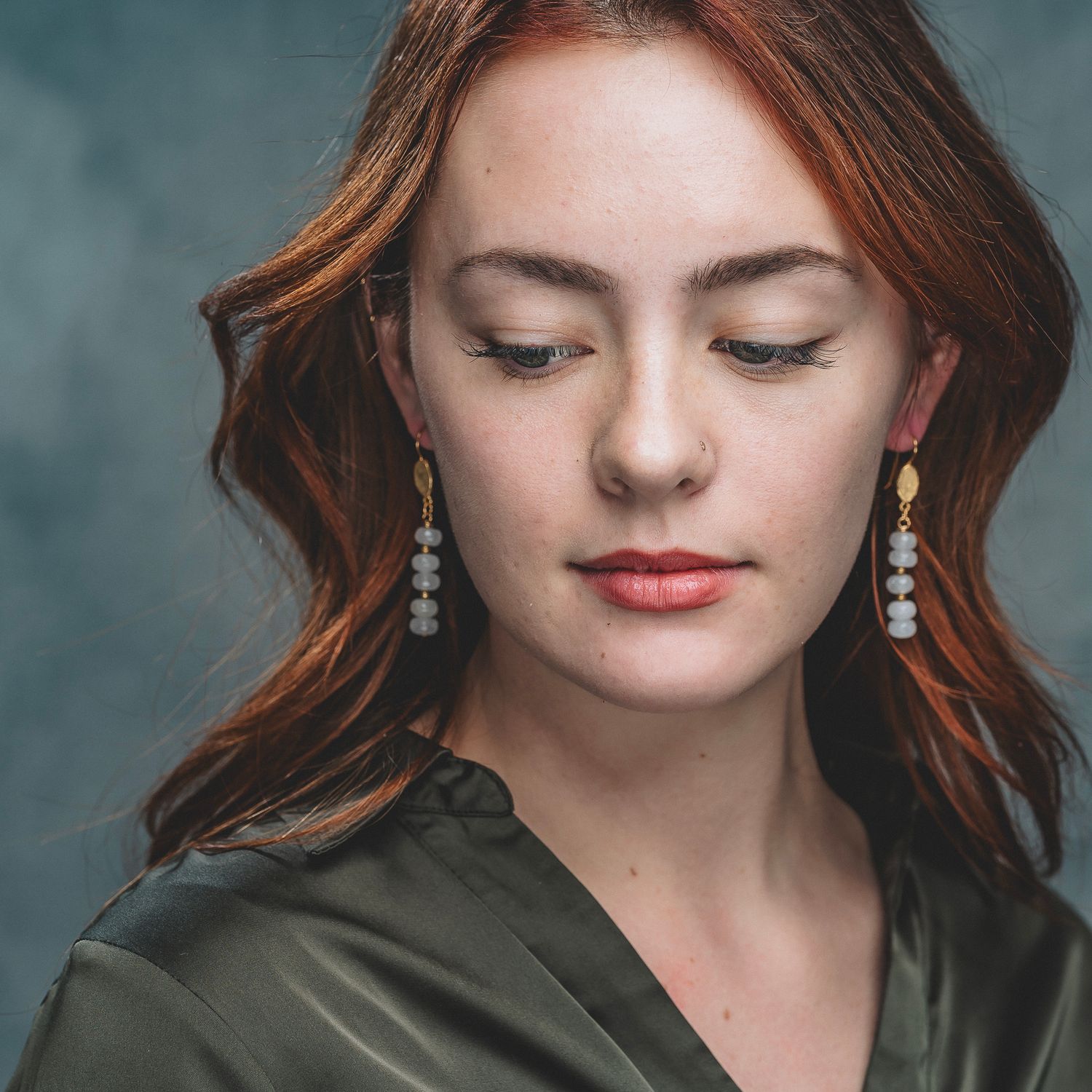 Woman with red wavy hair wearing elegant dangling pearl earrings and olive satin blouse.