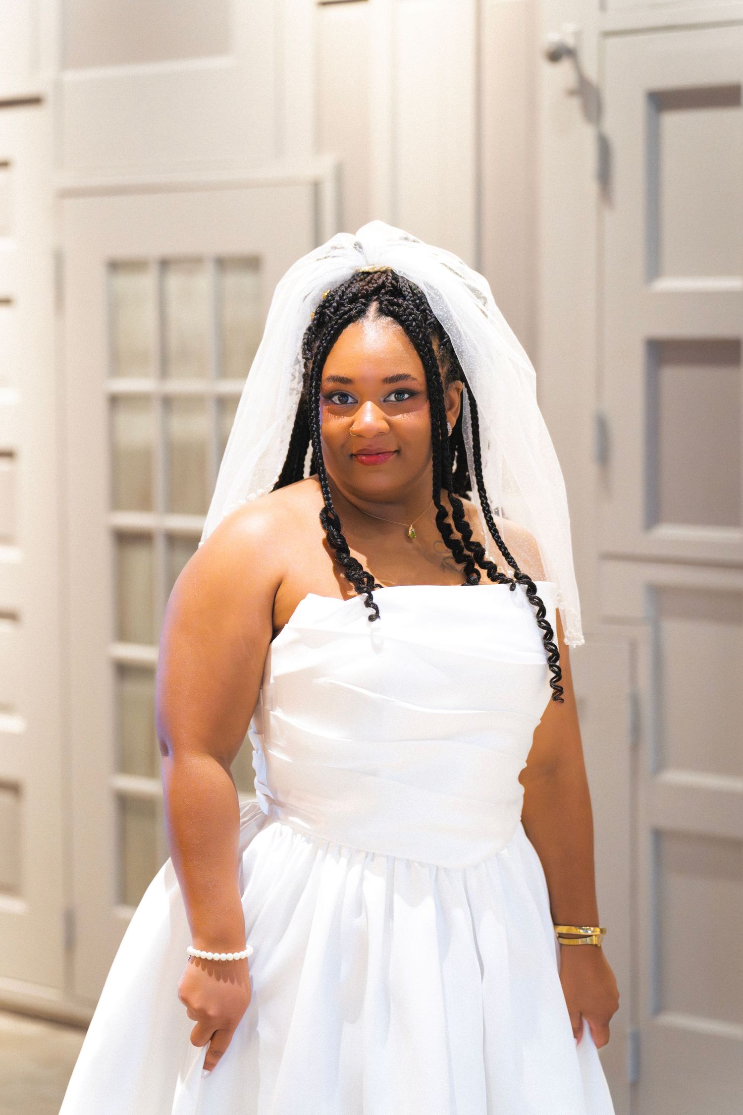 Black bride in strapless white gown and veil with braided hair posing indoors.