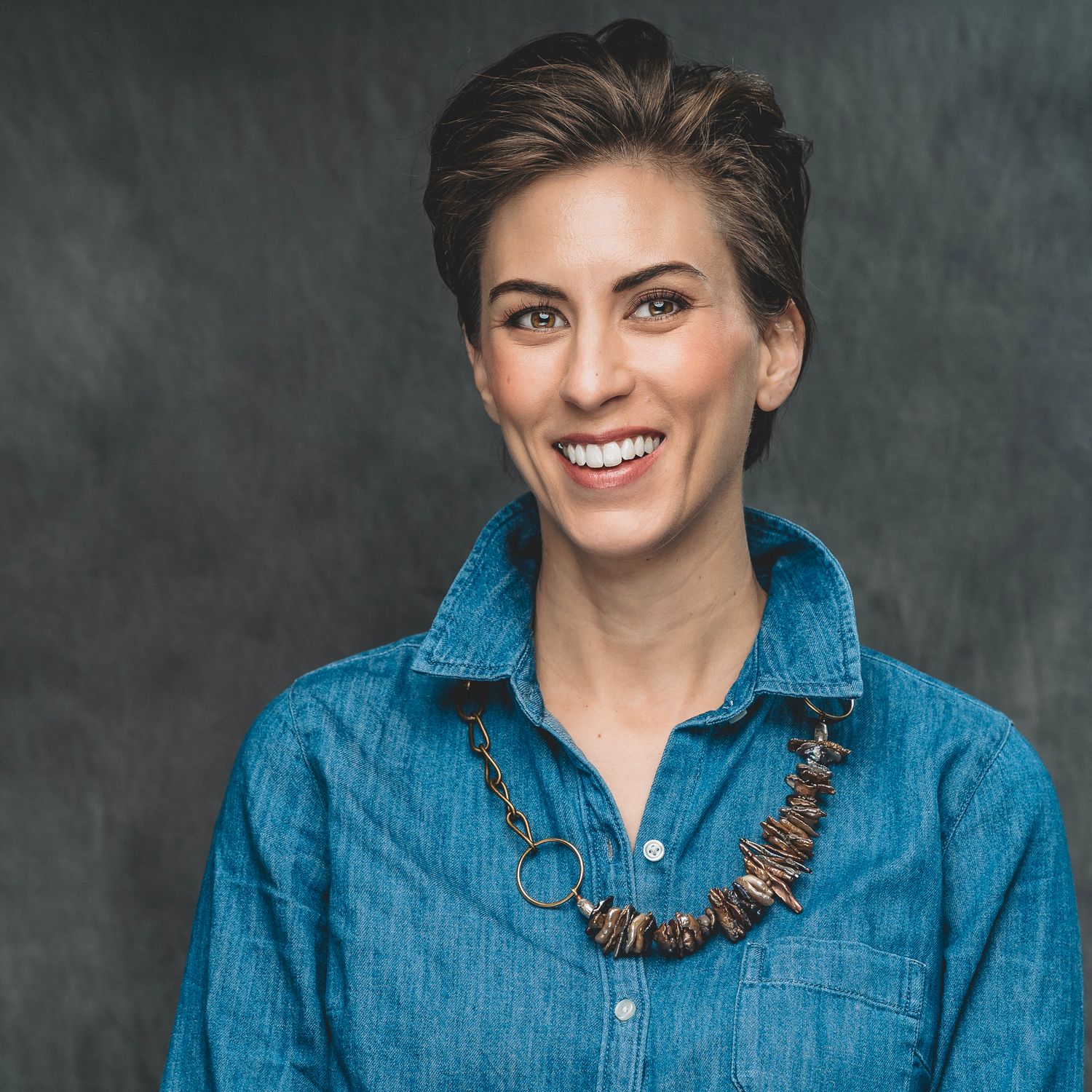 Smiling woman with short hair wearing a denim shirt and wooden necklace in a studio portrait.