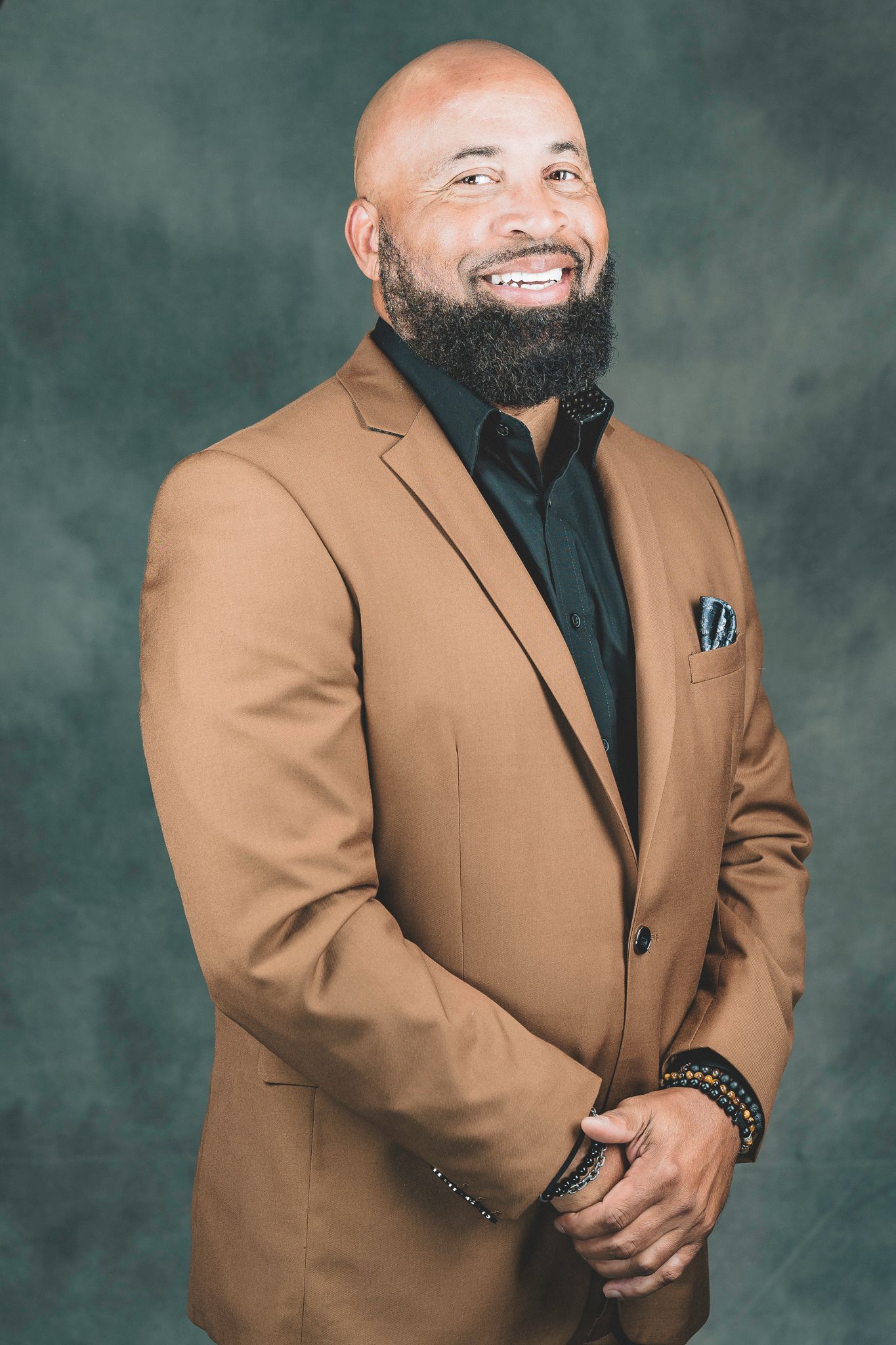 Smiling bald man with beard wearing a tan blazer and black shirt in a professional portrait.