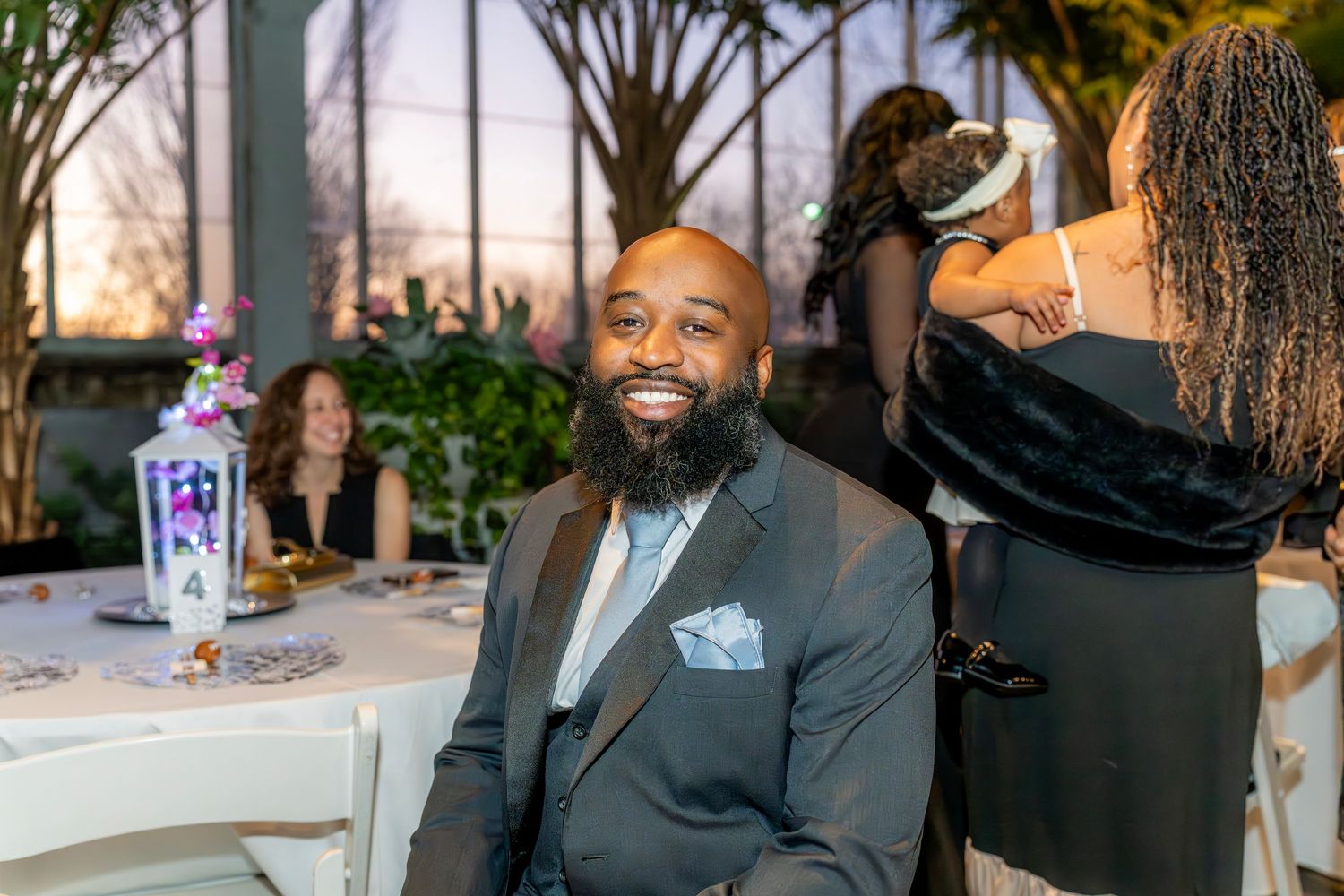 Bearded man in black suit smiling at wedding reception with guests in background.