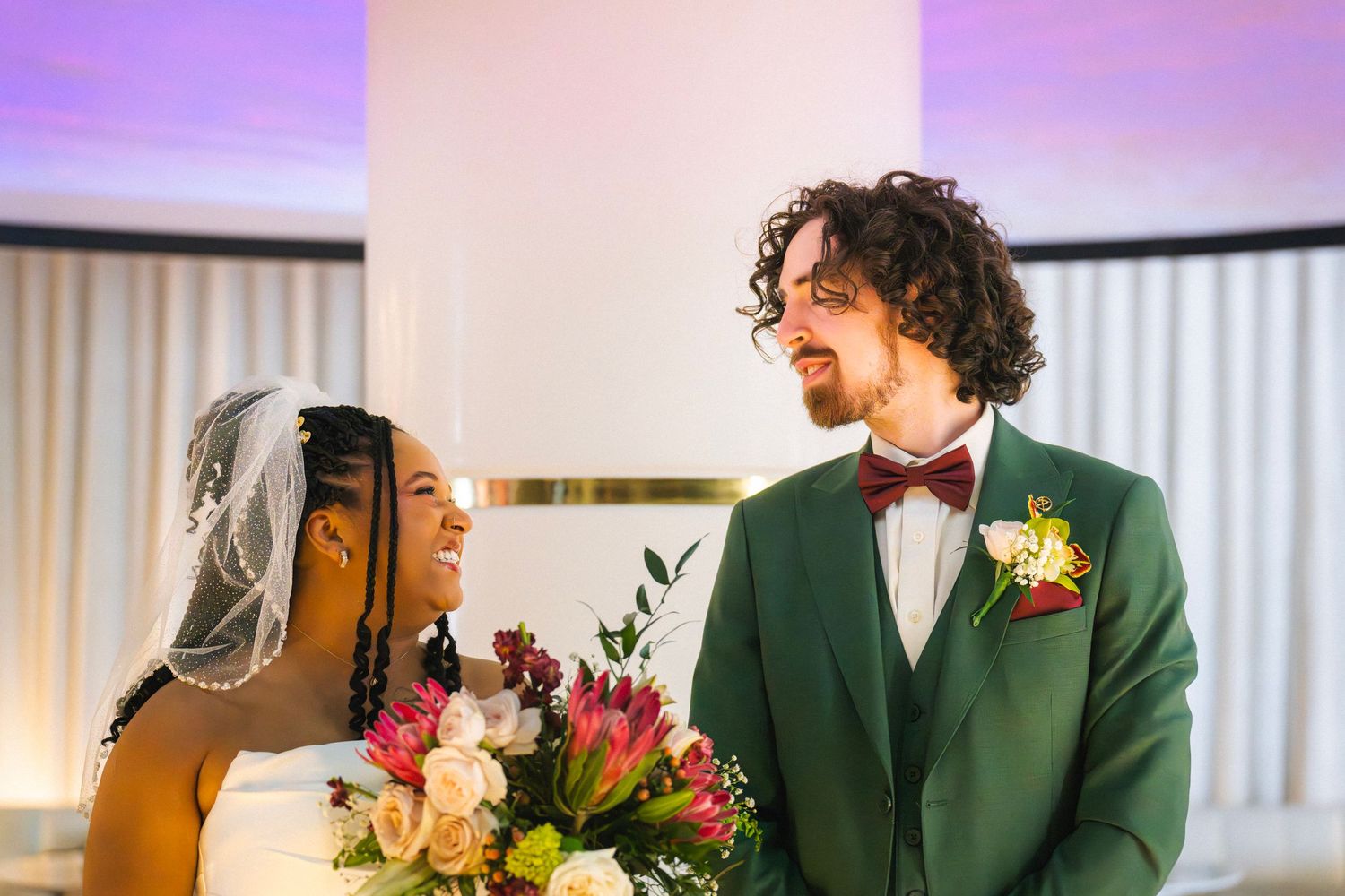 Interracial couple sharing a joyful moment at their wedding, bride in white veil holding colorful bouquet, groom in green suit.