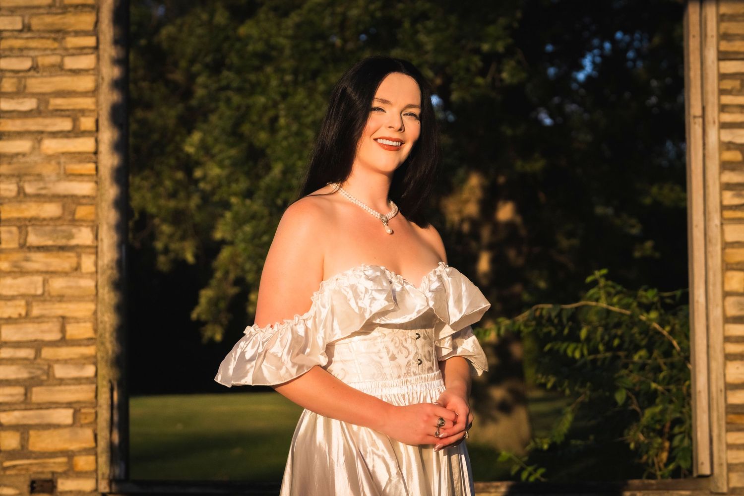 Smiling woman in elegant white off-shoulder dress bathed in golden sunset light outdoors.