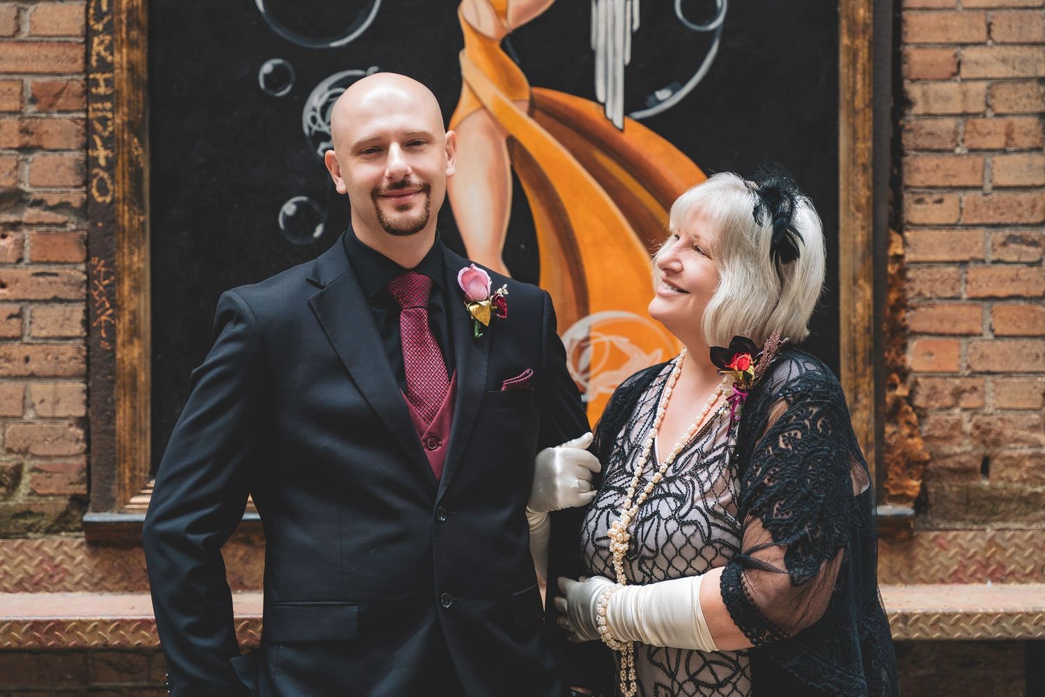 Bald groom in dark suit with burgundy tie poses with older woman in lace dress.