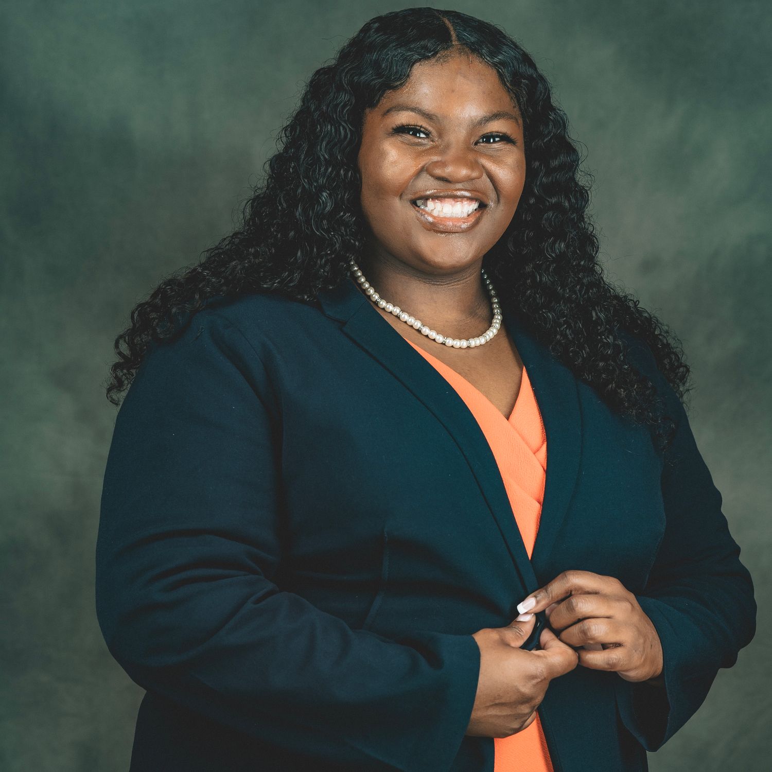 Professional headshot of a smiling woman in a navy blazer and orange top wearing a pearl necklace.