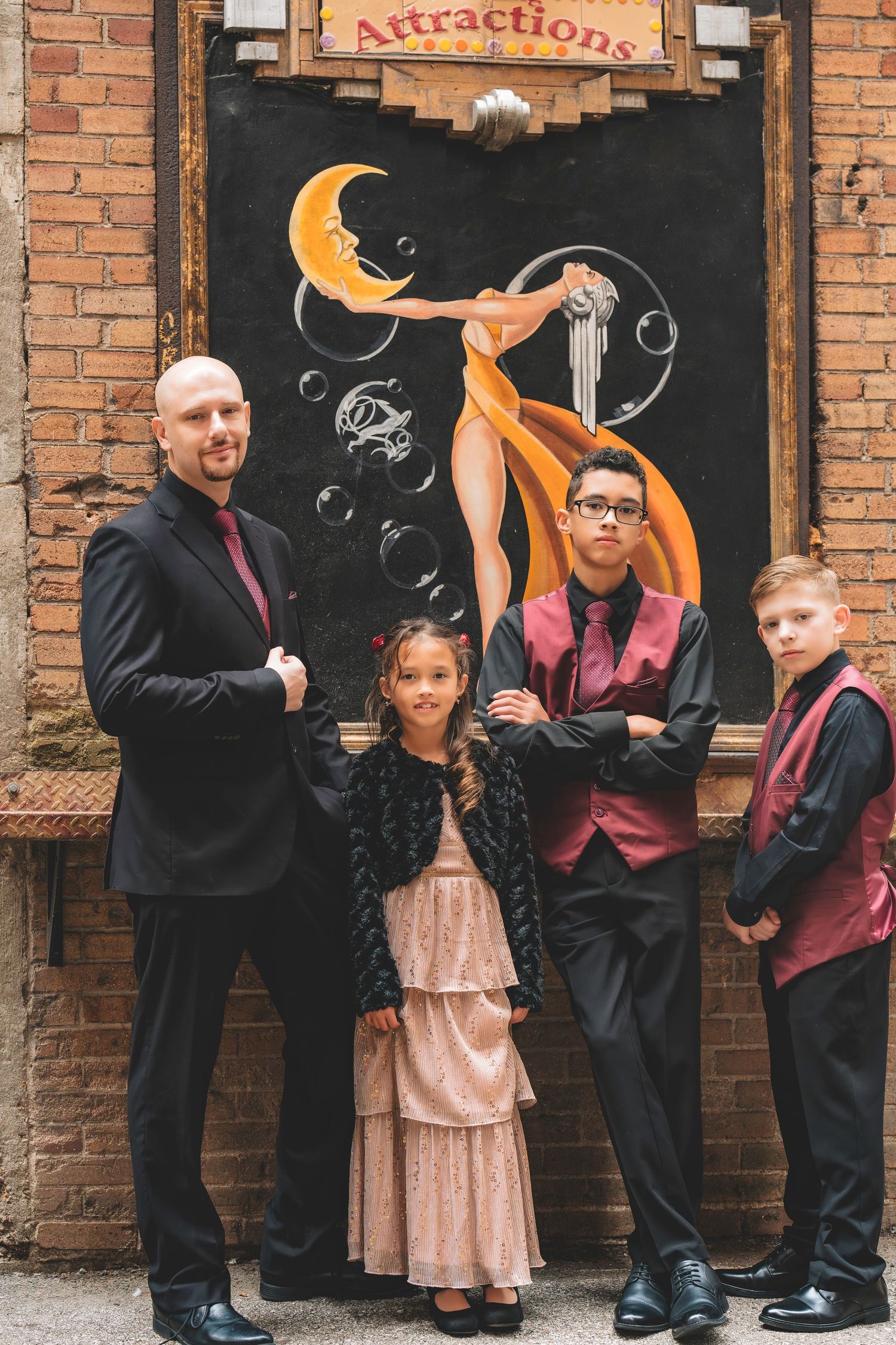 A man in a black suit poses with three children in formal attire in front of a colorful mural.