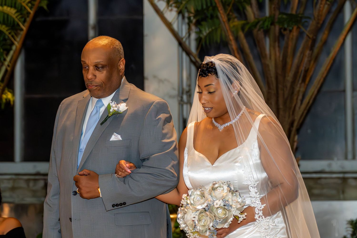 Father escorts bride in white wedding gown and veil down the aisle at outdoor ceremony.