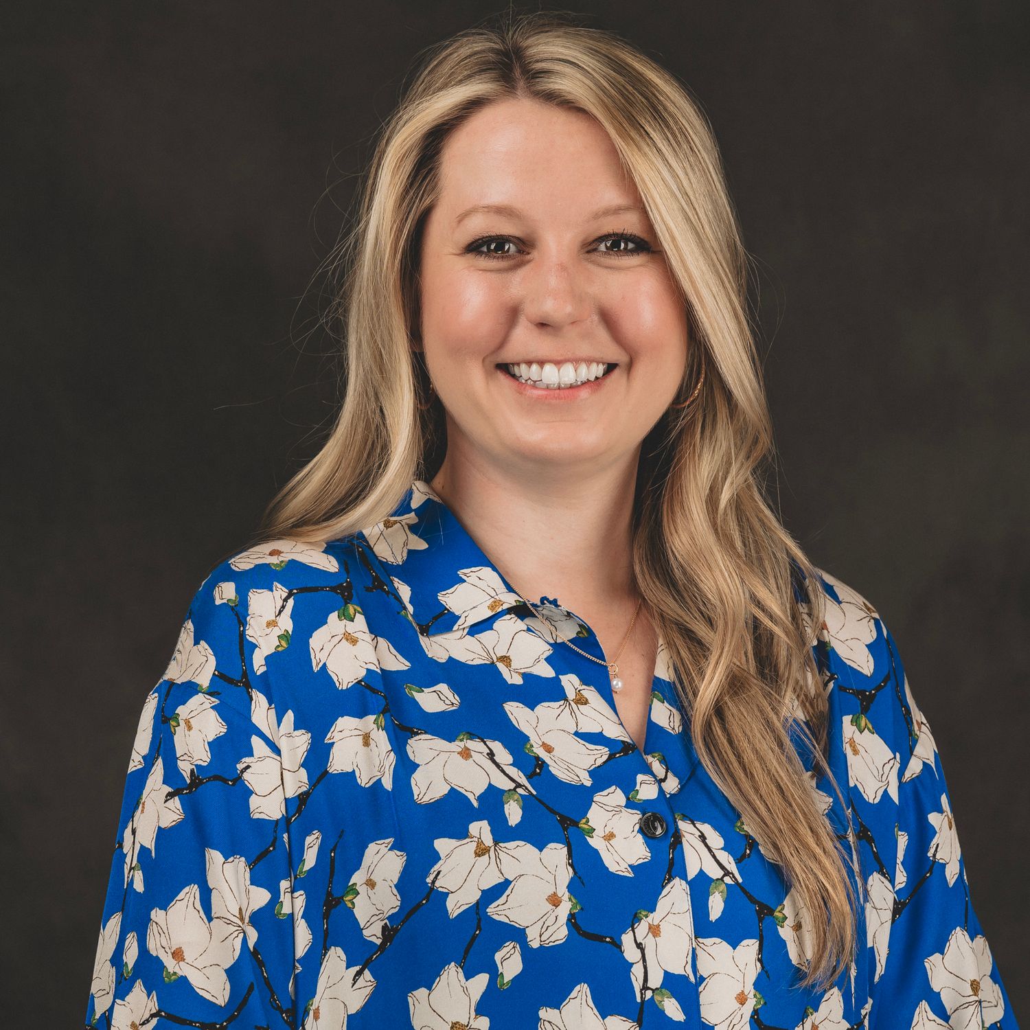 Professional headshot of a blonde woman smiling in a blue floral blouse against a dark background.