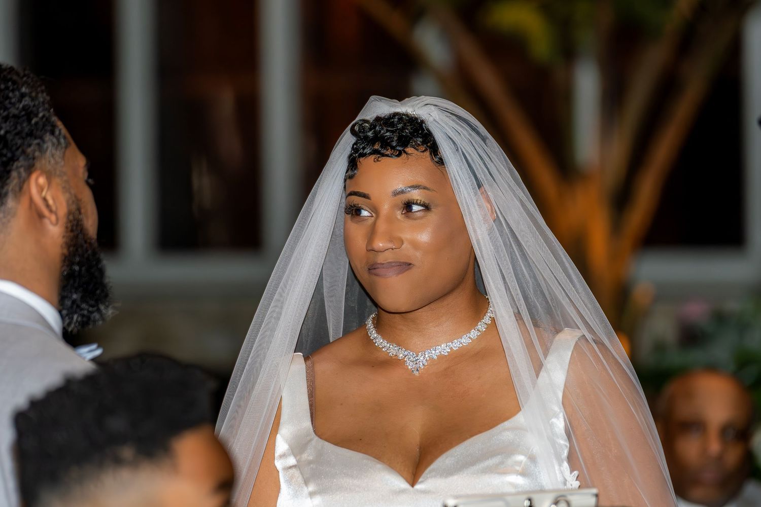 Beautiful bride in white gown and veil with diamond necklace gazes forward at wedding ceremony.