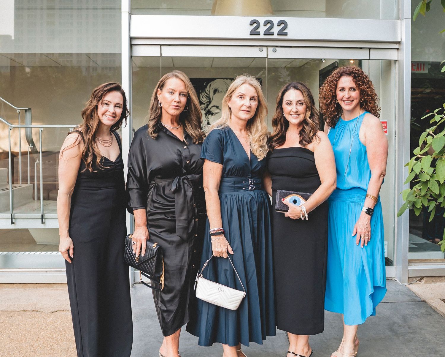 Five women in elegant dresses pose together outside a building numbered 222.