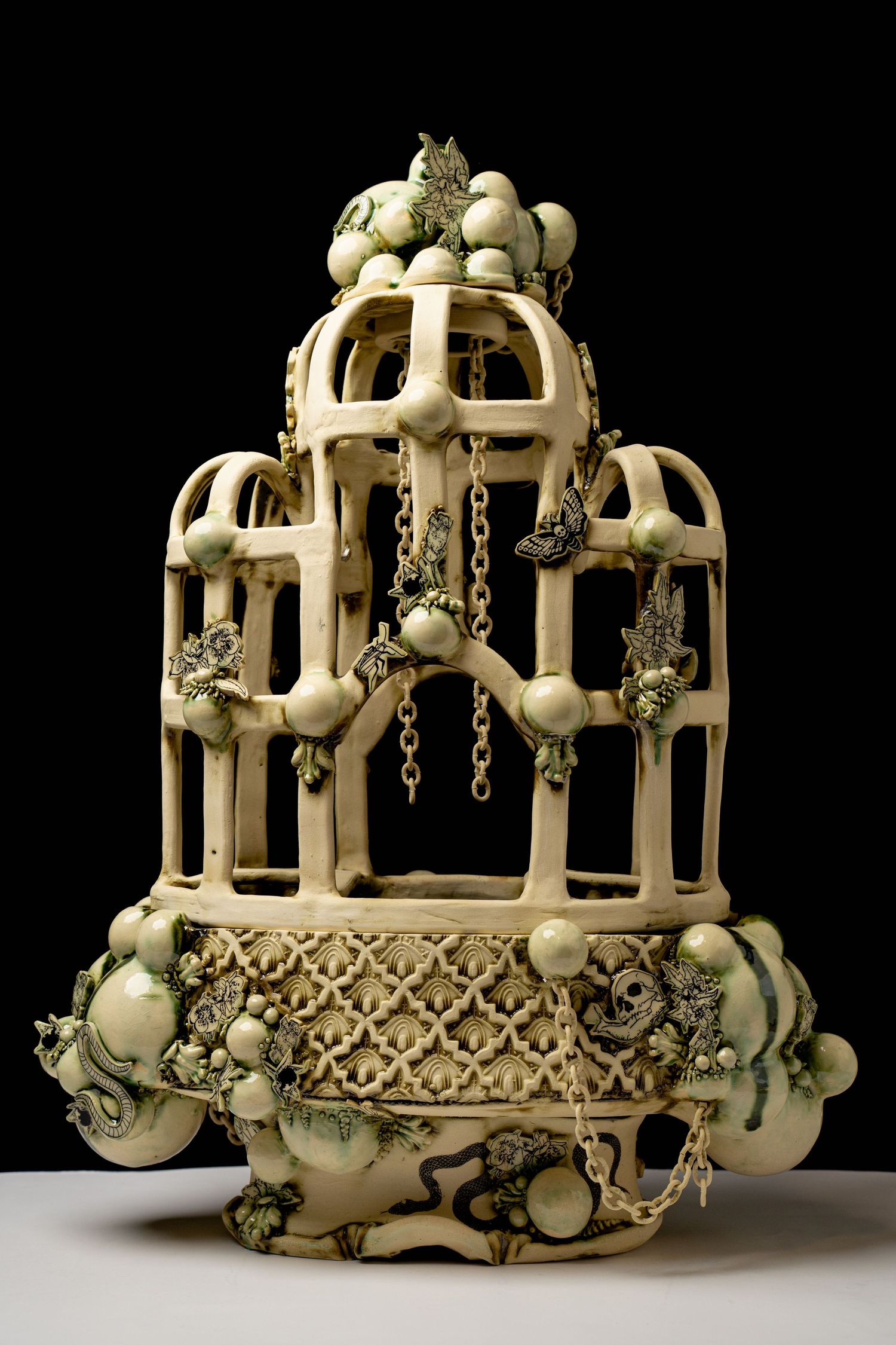 Ornate ceramic birdcage sculpture with green floral details and chains on dark background.