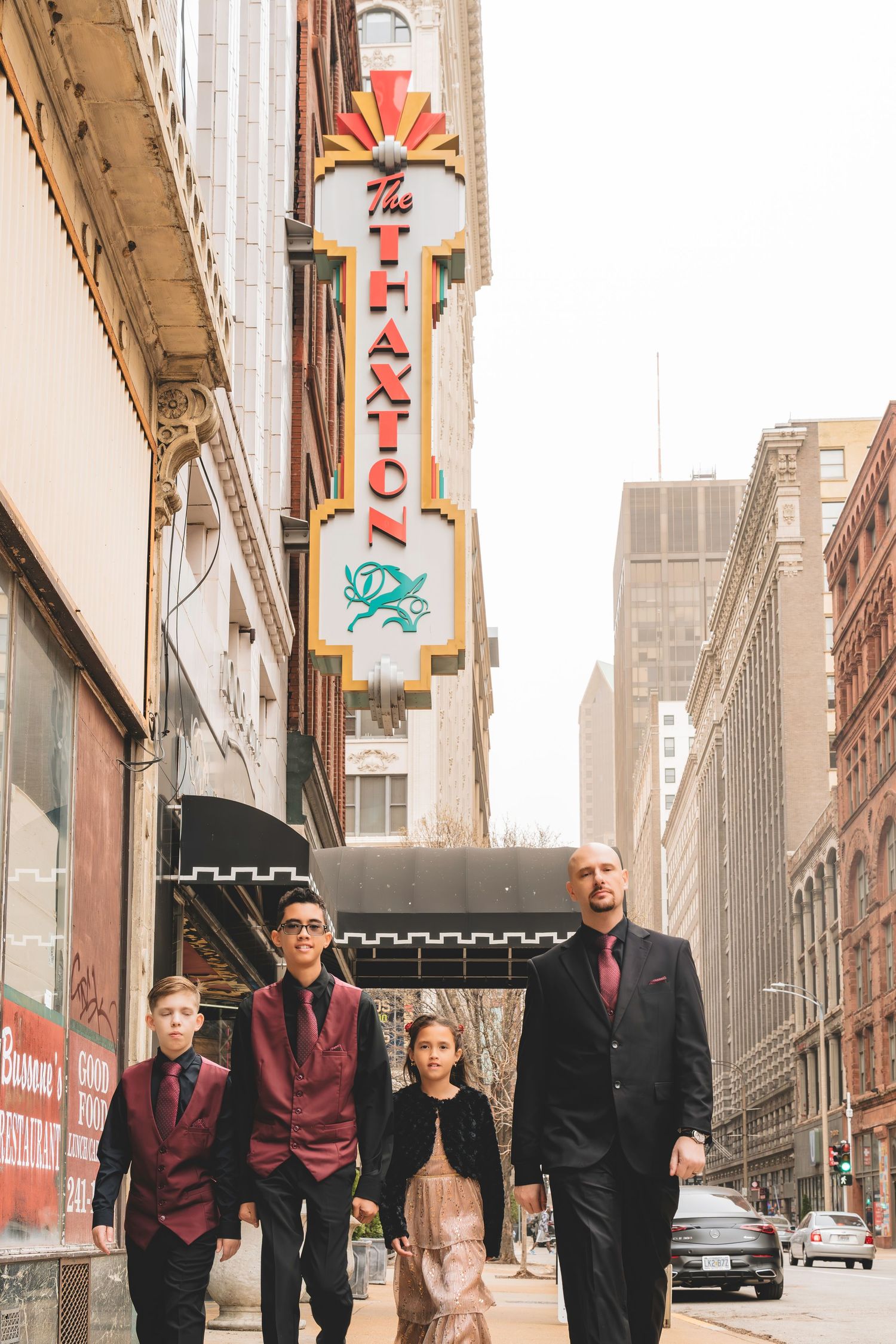 A father in a black suit walks with three children in formal wear under The Haxton theater sign.