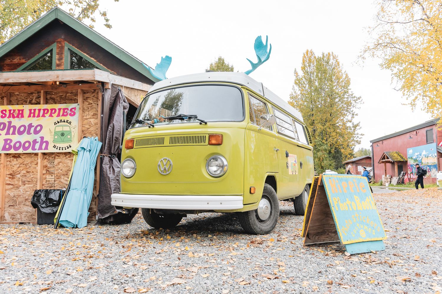 Vintage yellow VW camper van parked near a rustic photo booth sign at an outdoor autumn event.