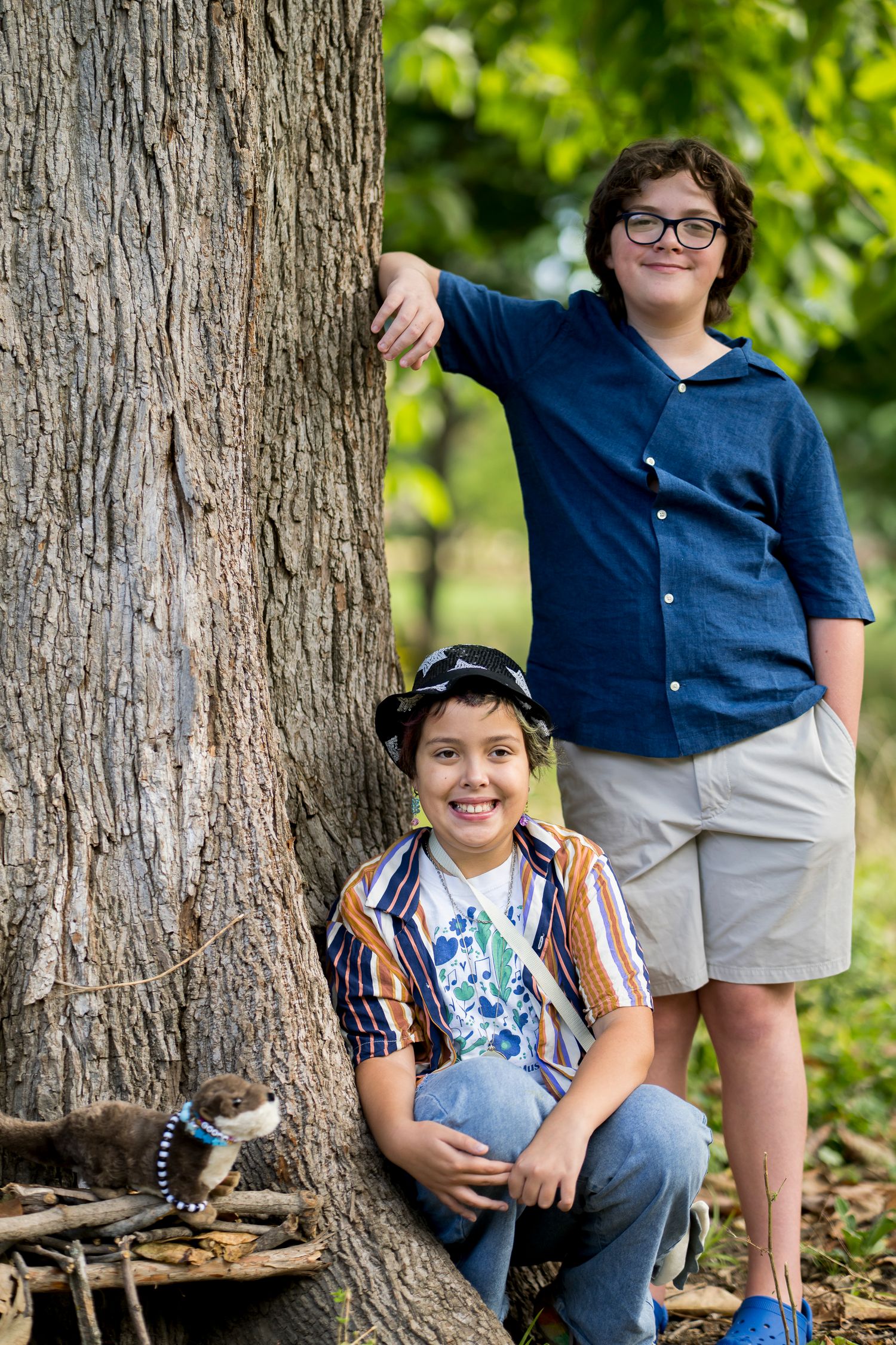 Two smiling boys standing by a large tree in a green outdoor park setting.
