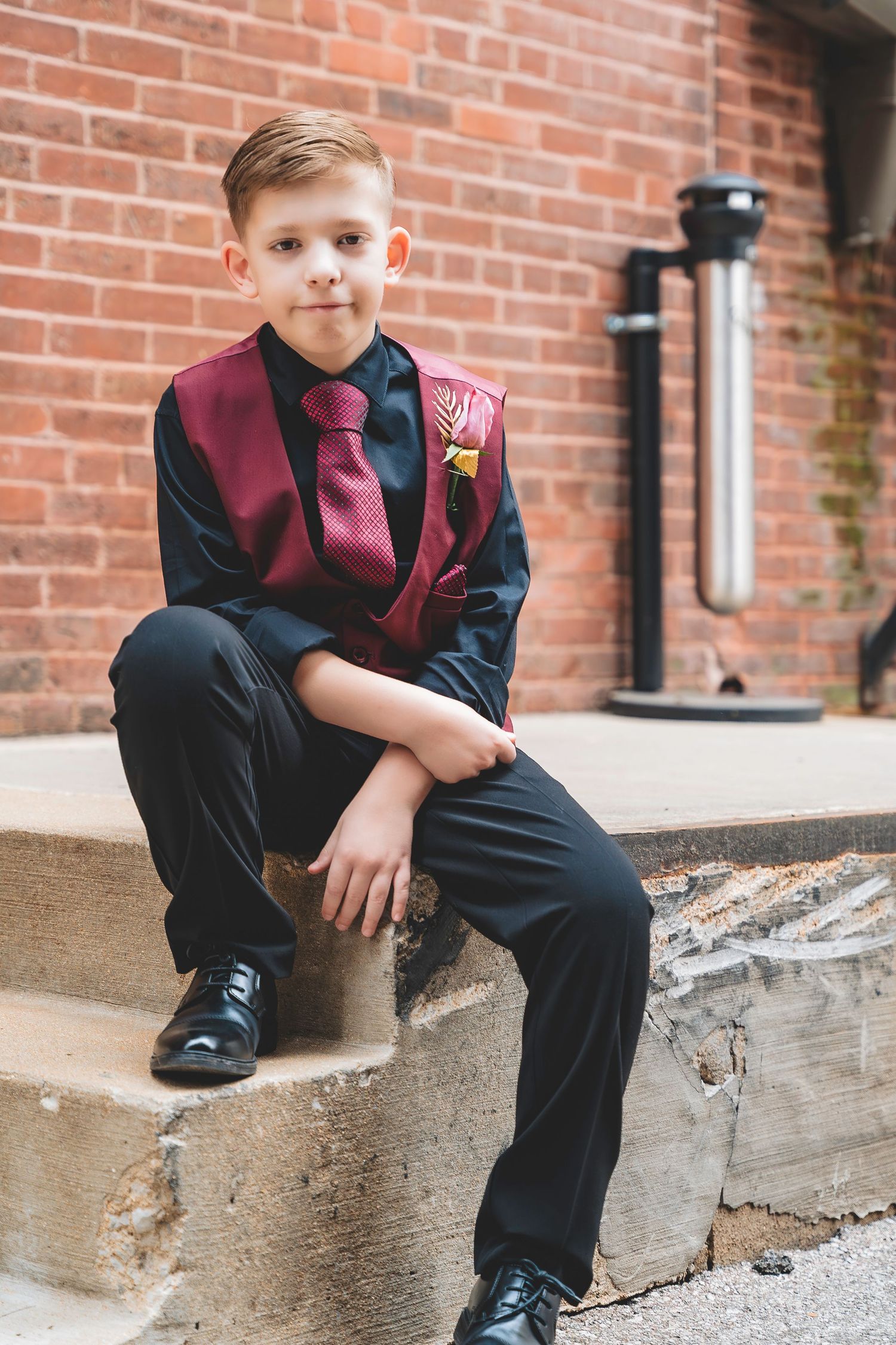 Young boy in burgundy vest and black suit sits on concrete steps near brick wall.