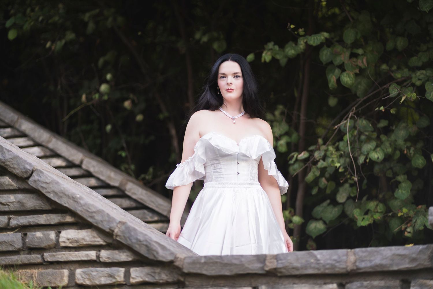 Dark-haired woman in white off-shoulder dress standing on stone steps surrounded by green trees.