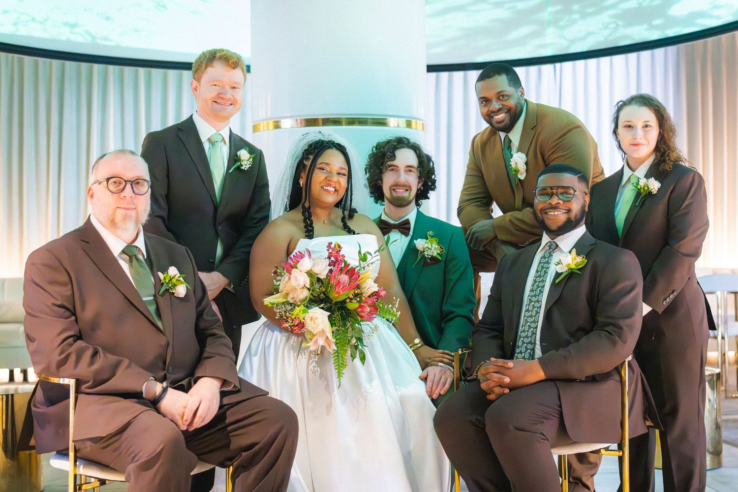 Wedding party group photo with bride in white gown, groom in green suit, groomsmen in brown suits, indoors with elegant decor.