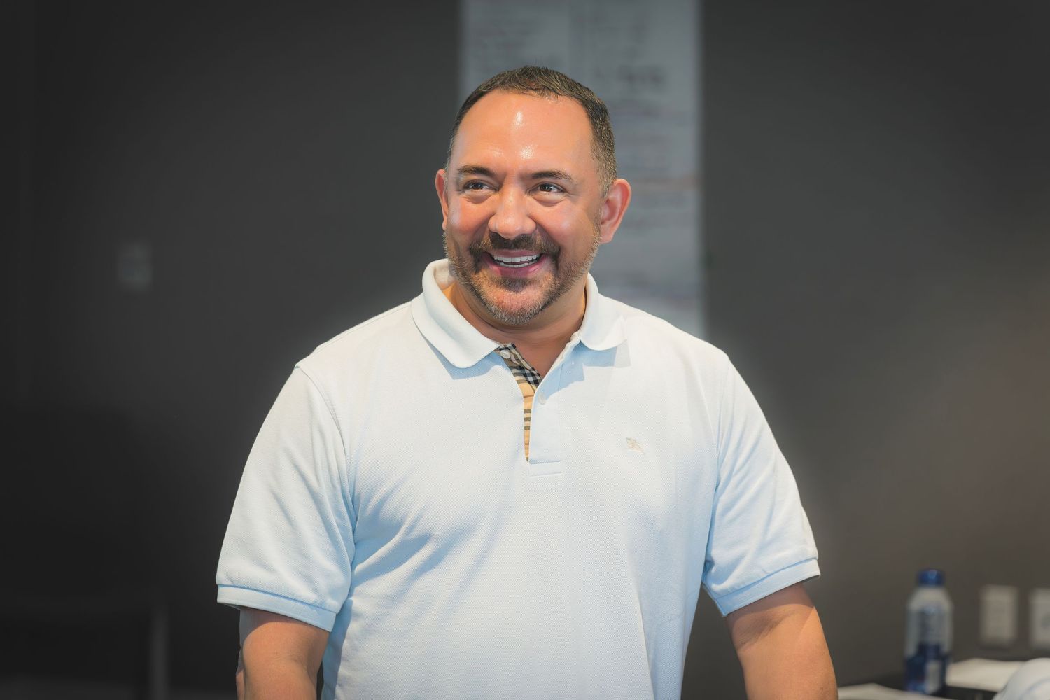 Smiling man in light blue polo shirt standing in a modern office or meeting room.