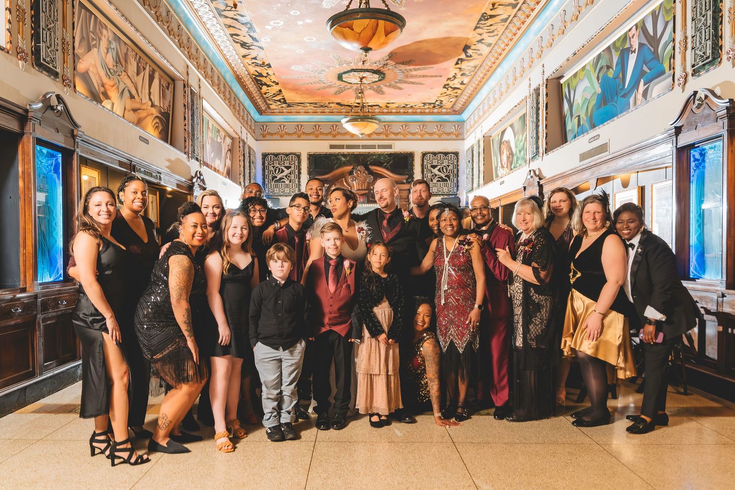 Large wedding party group photo in an ornate gilded hallway with colorful murals.