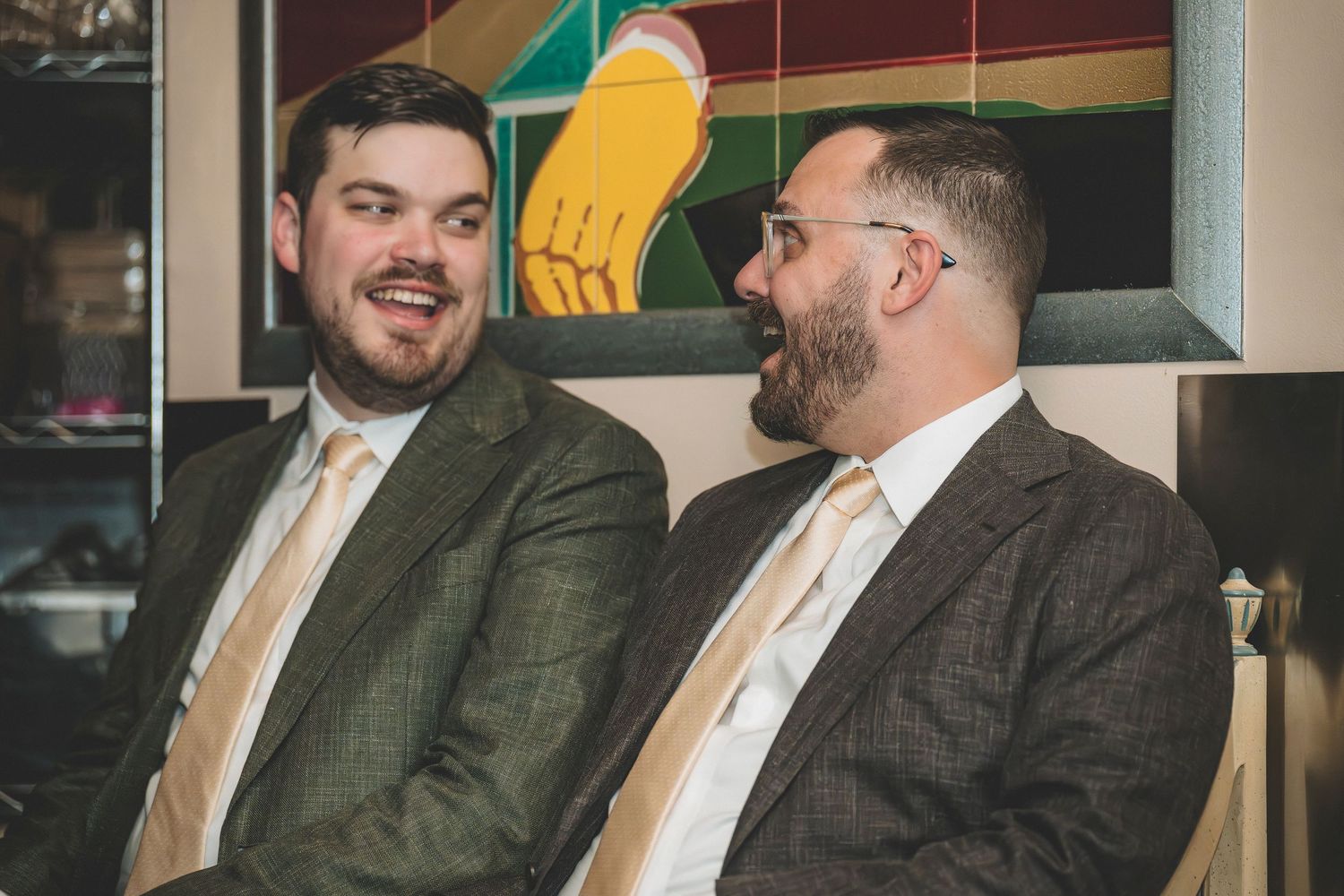 Two groomsmen in matching gray suits share a laugh while seated at a wedding reception venue.