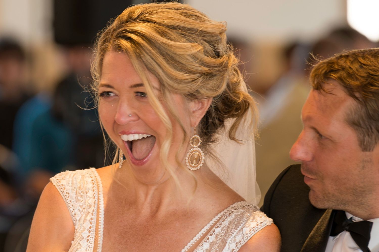 Blonde bride laughing joyfully wearing large earrings and lace wedding gown at reception.