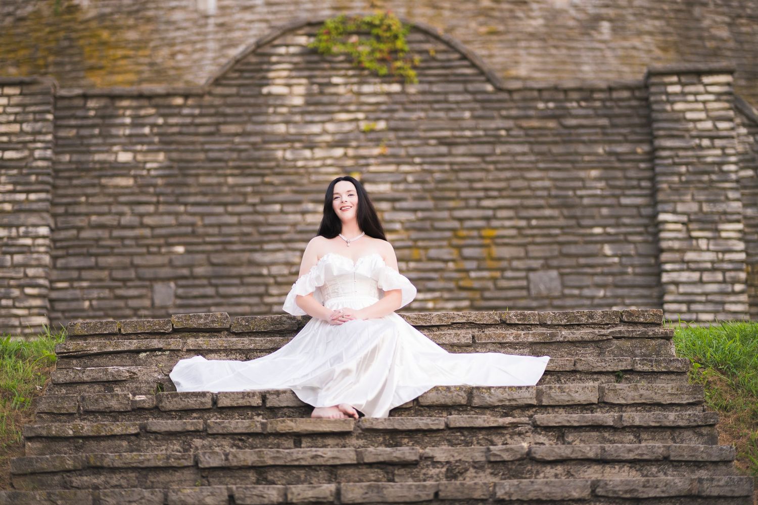 Pregnant woman in flowing white dress sitting gracefully on stone steps at outdoor venue.