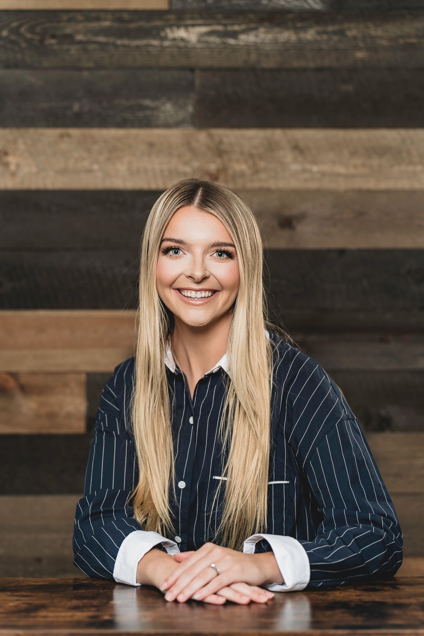 Smiling young blonde woman with long hair in striped shirt seated at wooden table with rustic wood wall background.