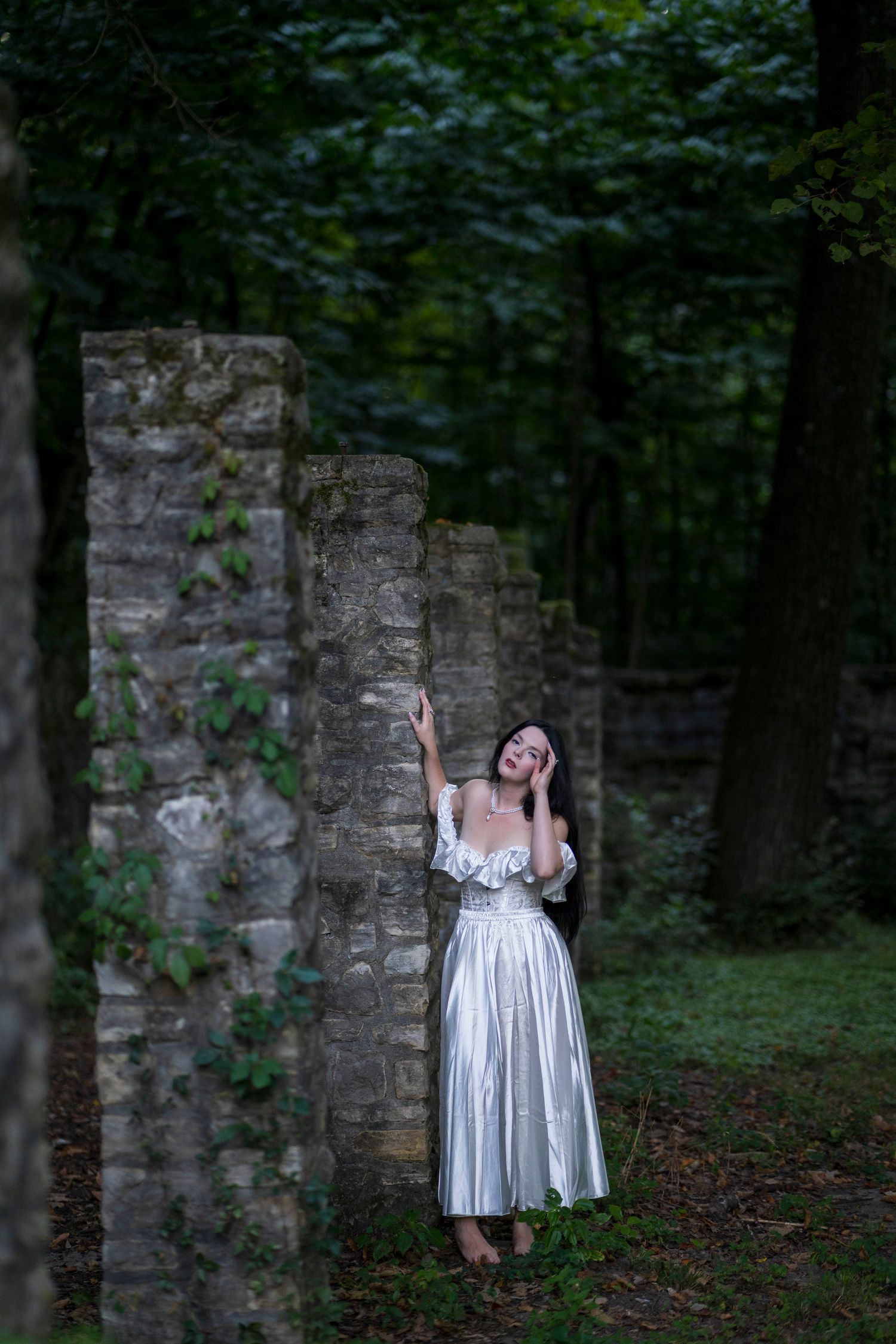 Woman in a silver white gown standing among stone pillars in a lush green forest setting.