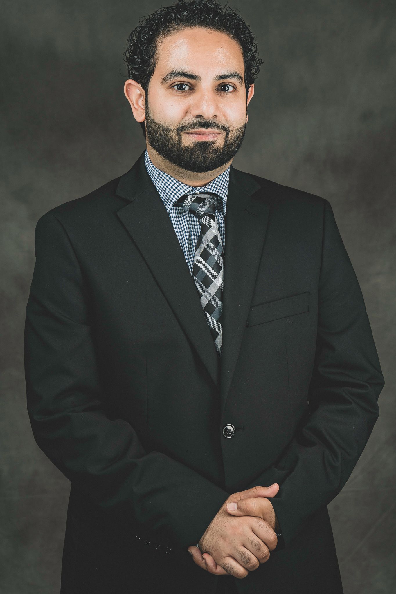 Professional man with beard in black suit and plaid tie standing with clasped hands against gray background.
