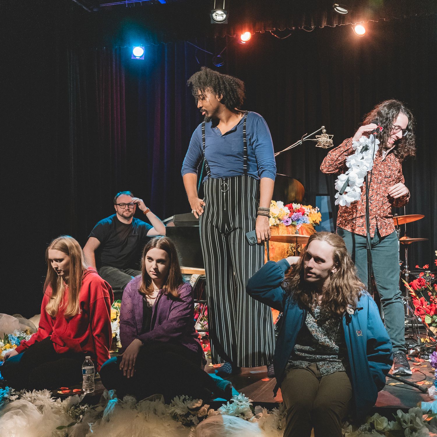 Diverse theater group performing on stage with colorful props under dramatic stage lighting.
