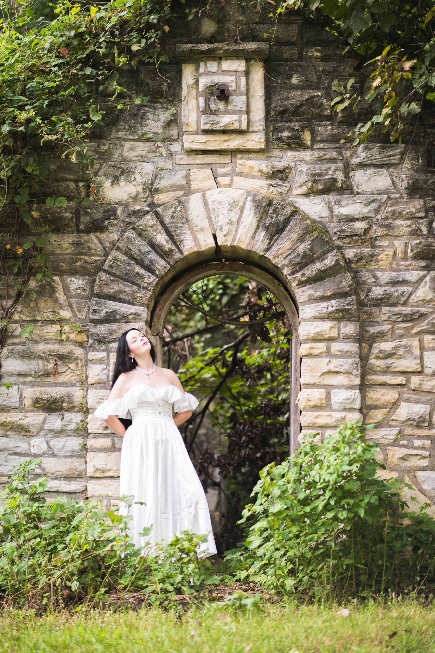 Woman in white off-shoulder dress standing in ivy-covered stone archway in garden setting.