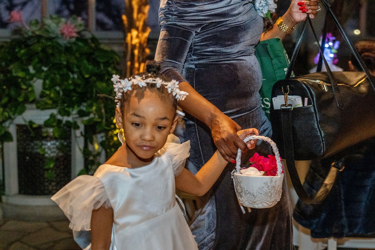 Adorable flower girl in white dress with floral crown holds basket at evening wedding reception.