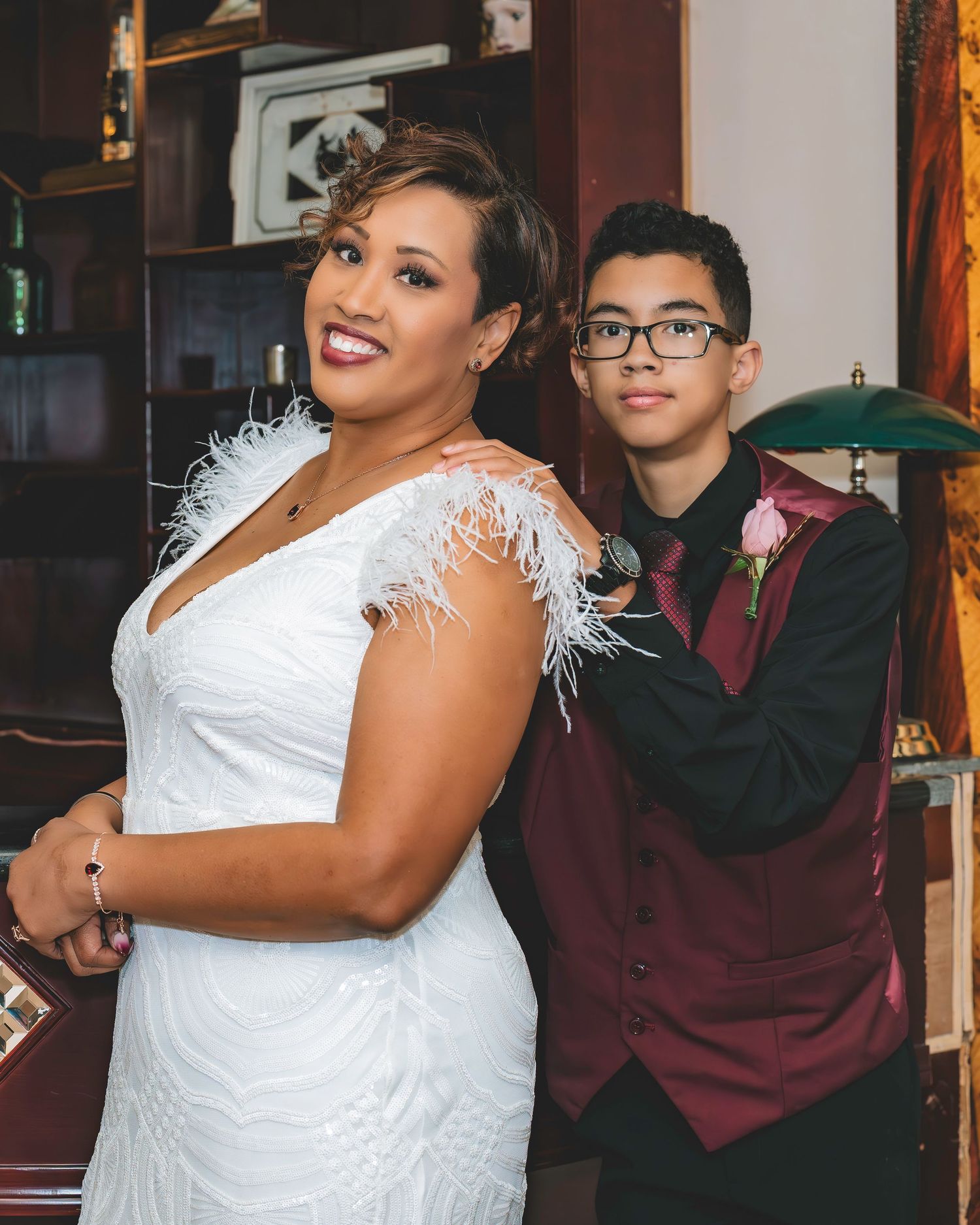 A bride in a white feathered gown poses with a teenage boy in a burgundy suit at a wedding.