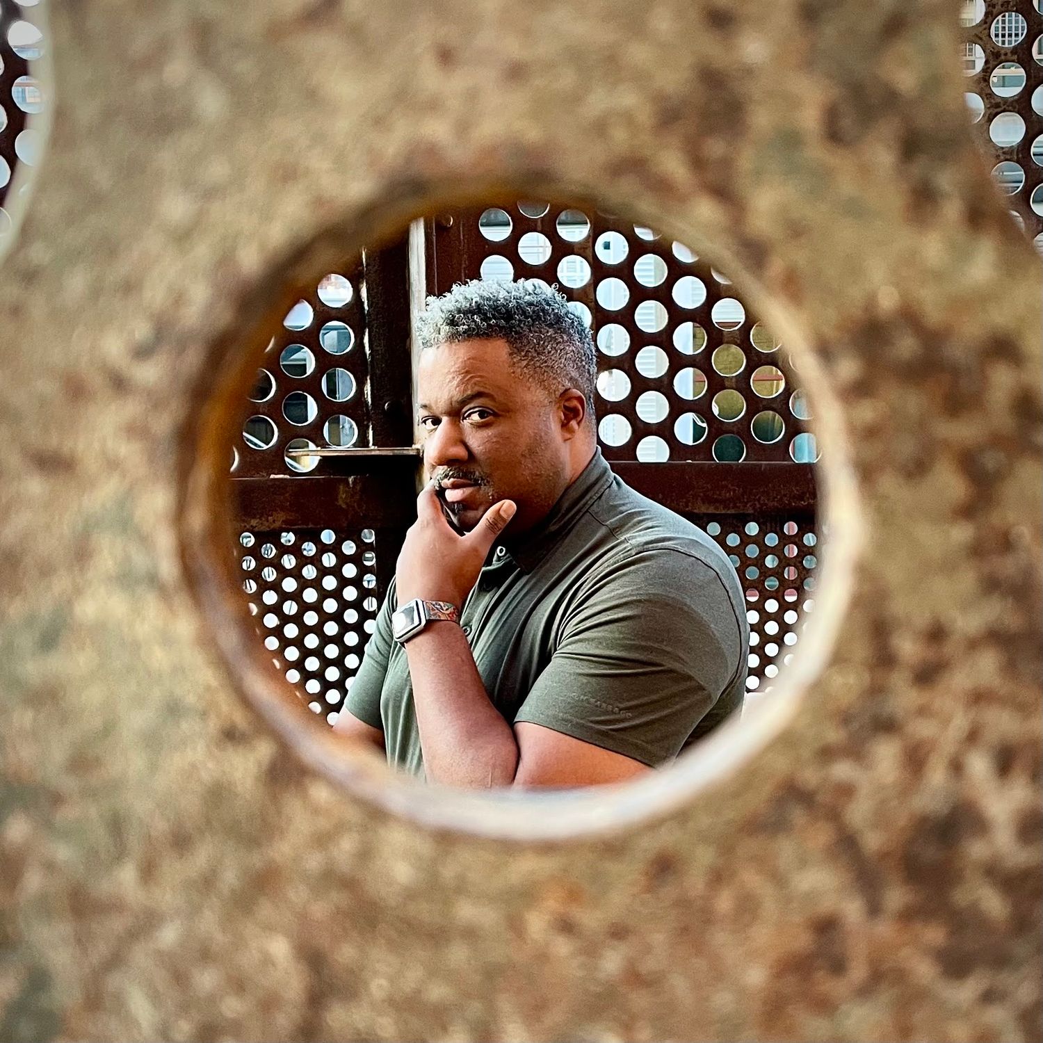Black man viewed through circular hole in rusty metal surface, thoughtful expression.