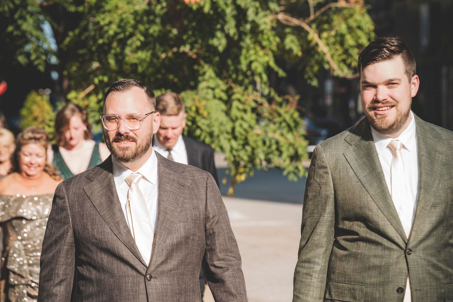 Two groomsmen in gray suits smile as they walk outdoors at a sunny wedding celebration.
