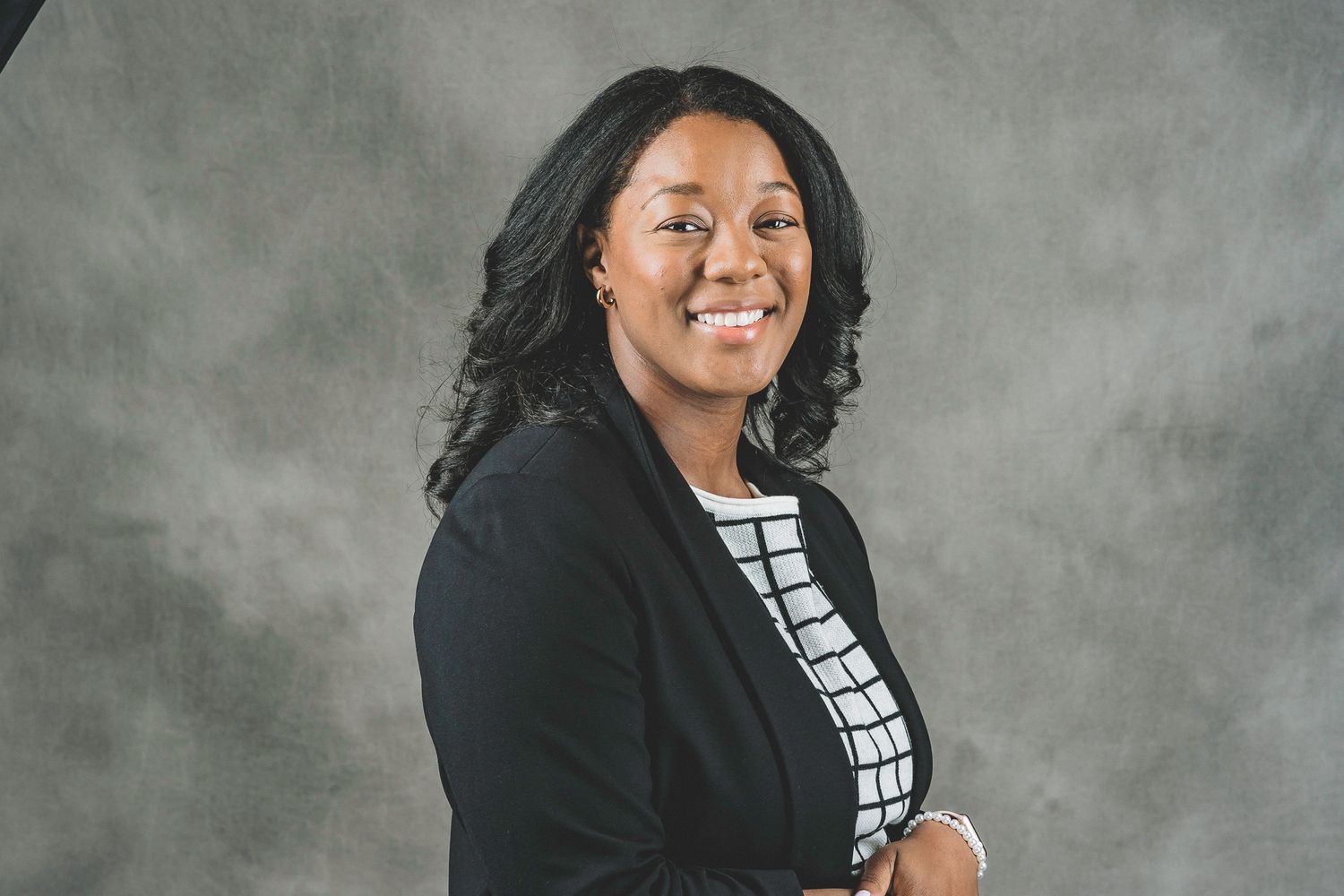 Smiling professional woman in black blazer and patterned top posing against a gray studio background.