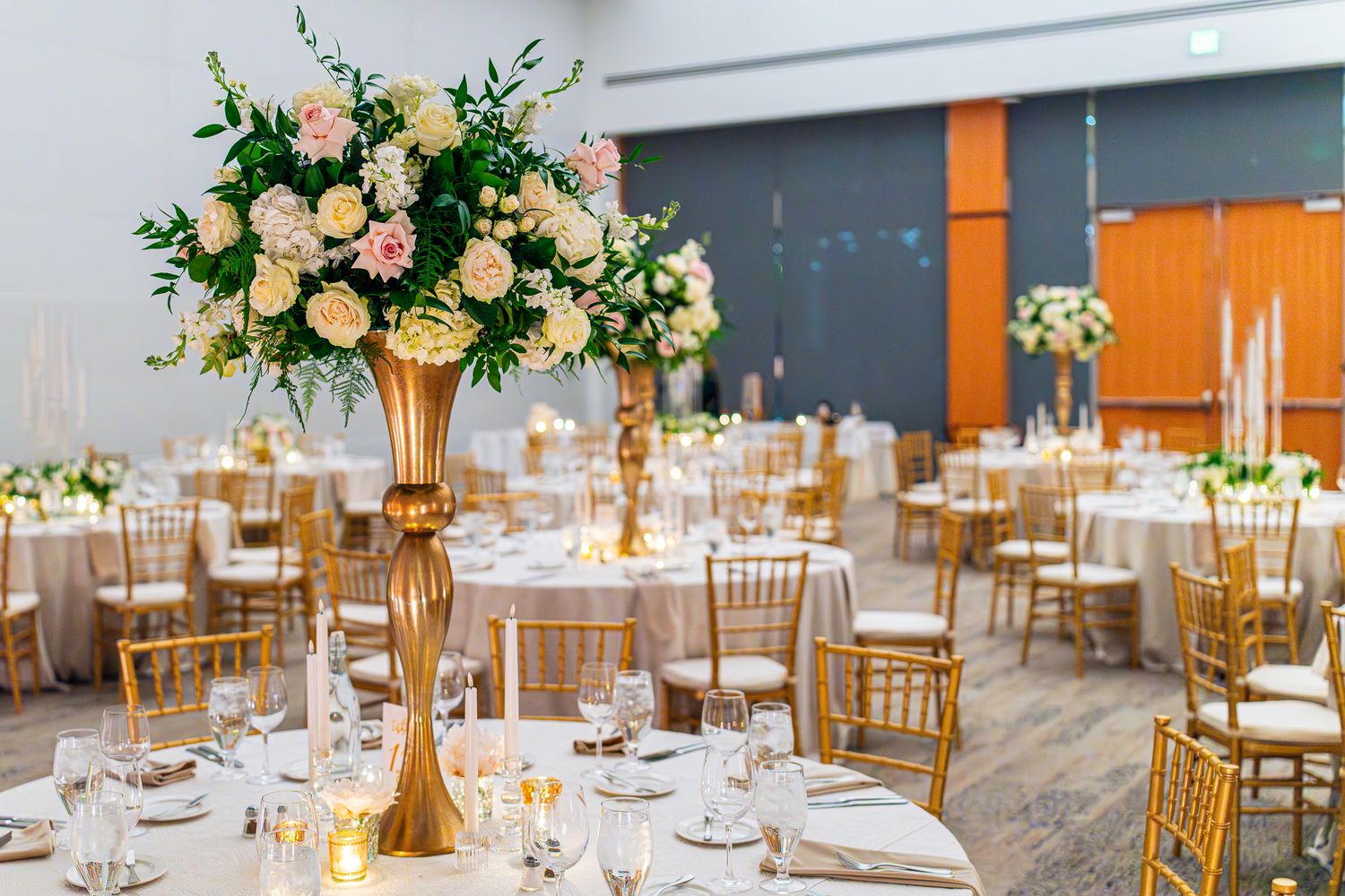 Elegant wedding reception hall with tall gold vases holding white and pink floral centerpieces on round tables.