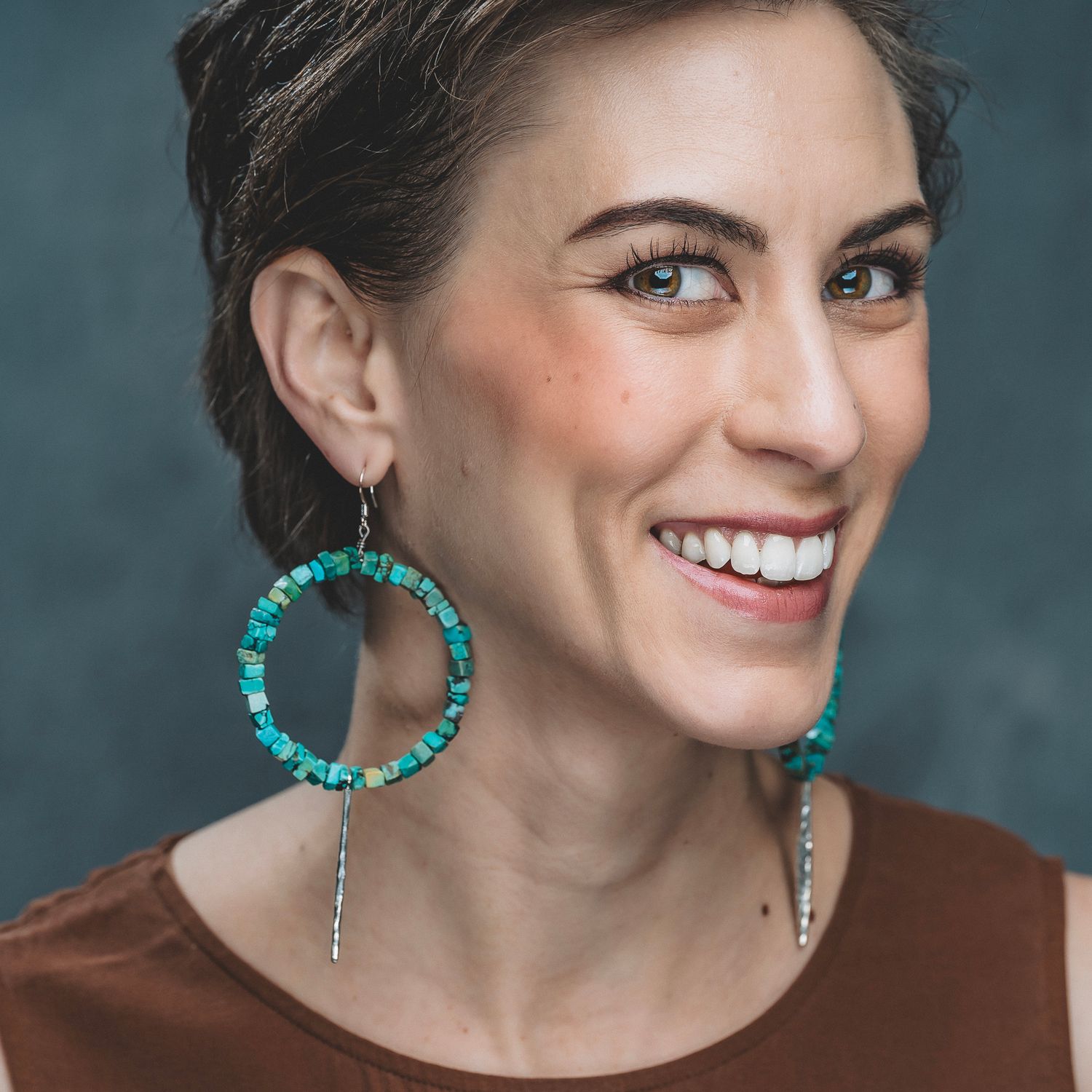 Smiling woman wearing large turquoise stone hoop earrings with silver chain accents.