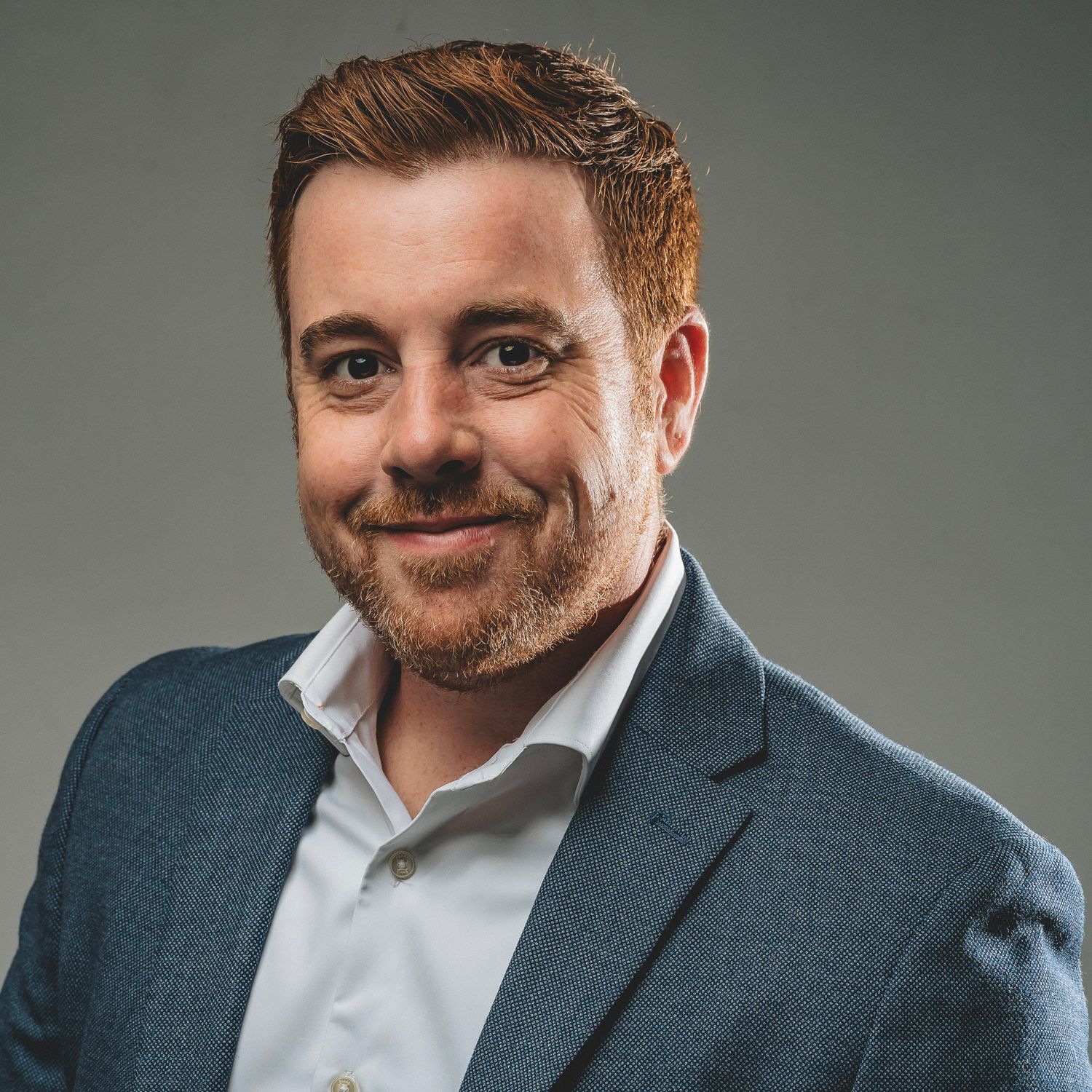 Smiling man with red hair wearing a blue blazer in a professional studio headshot.