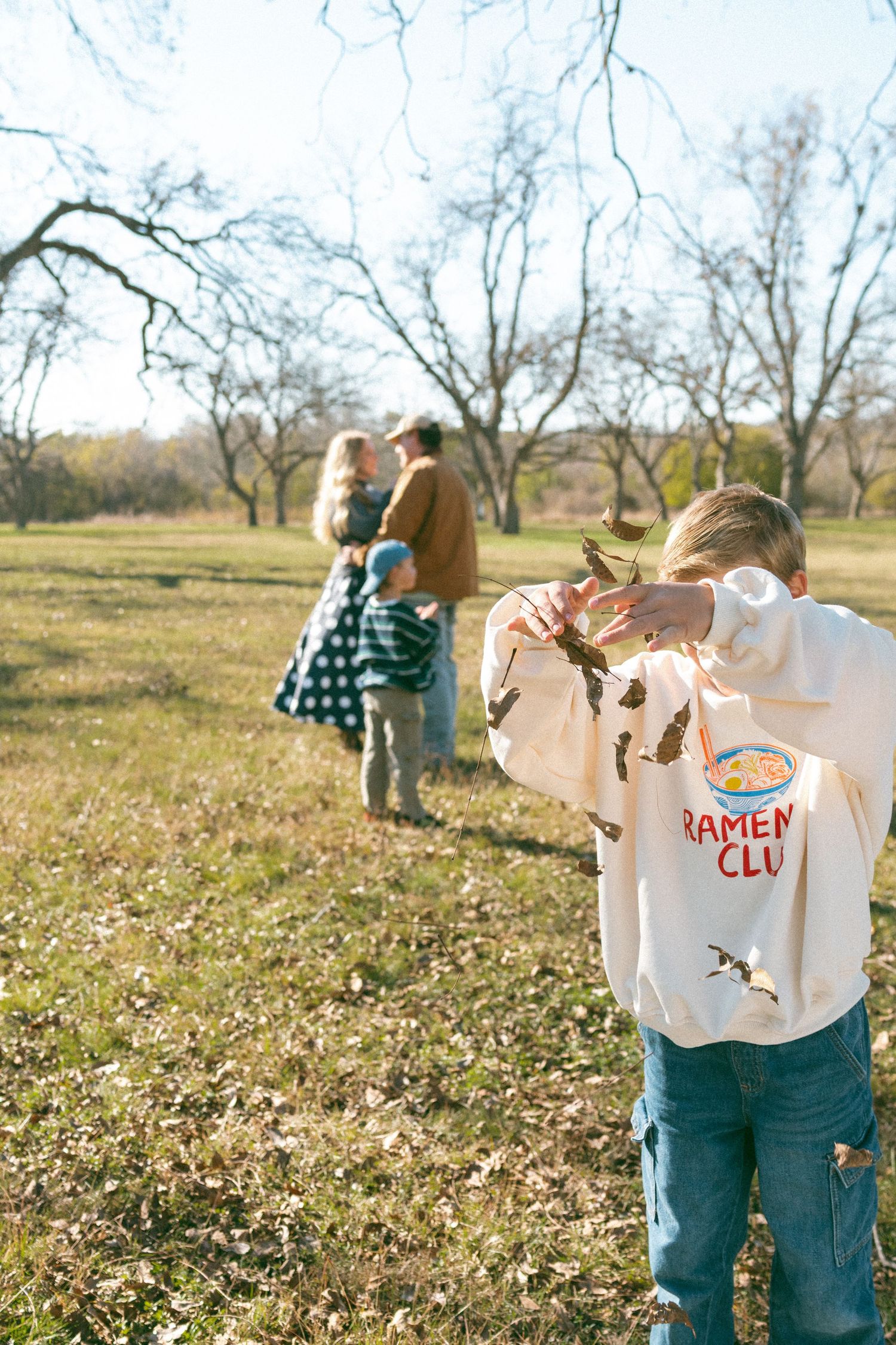 Children playing and running in a sunlit autumn field with fallen leaves and bare trees in the background.