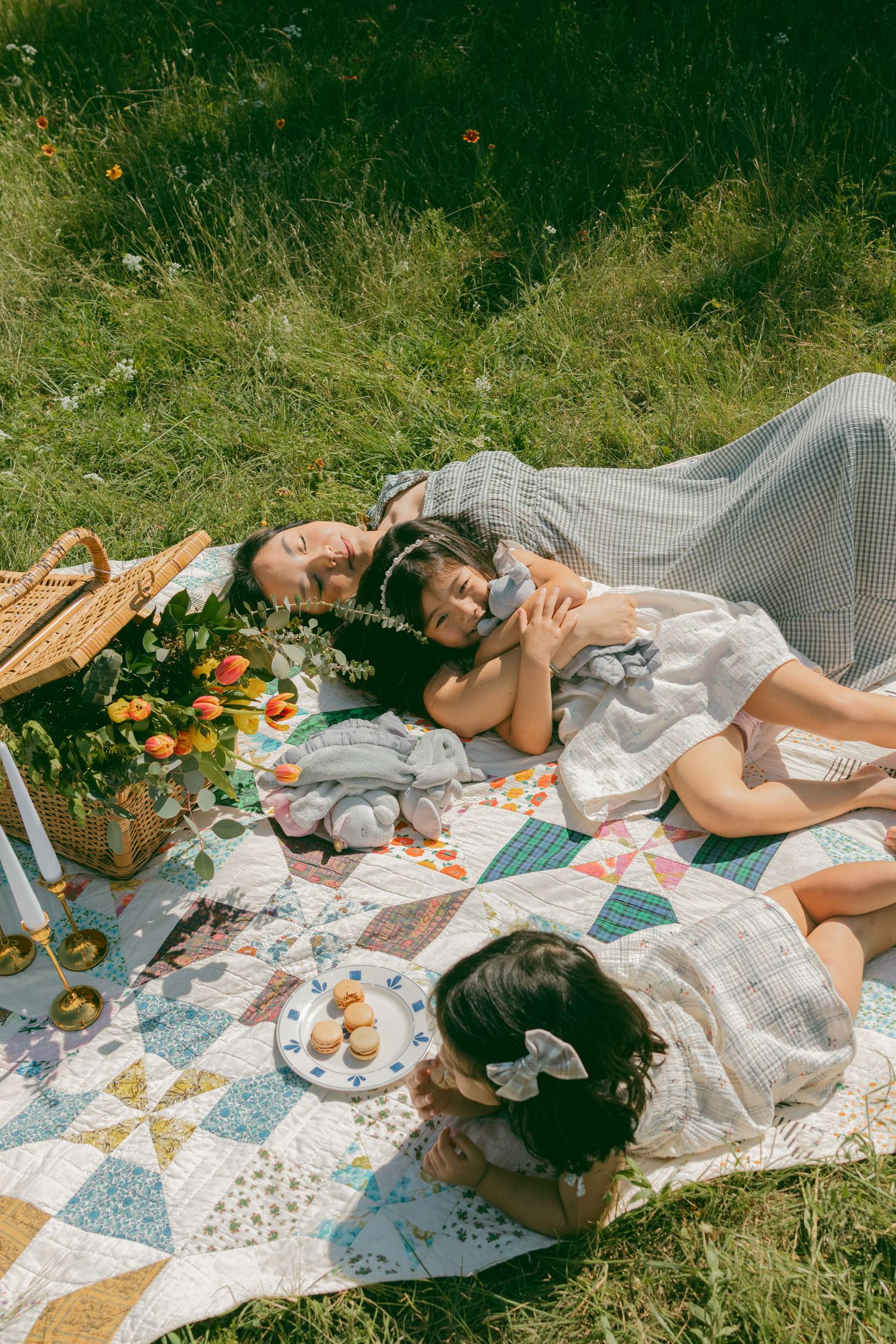 A dreamy picnic scene with a mother and two daughters, scattered food and blankets on grass in natural, direct light.