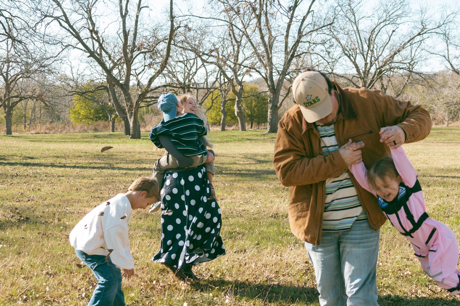Family members hold hands and dance together in a circle on a grassy field during a sunny autumn day.