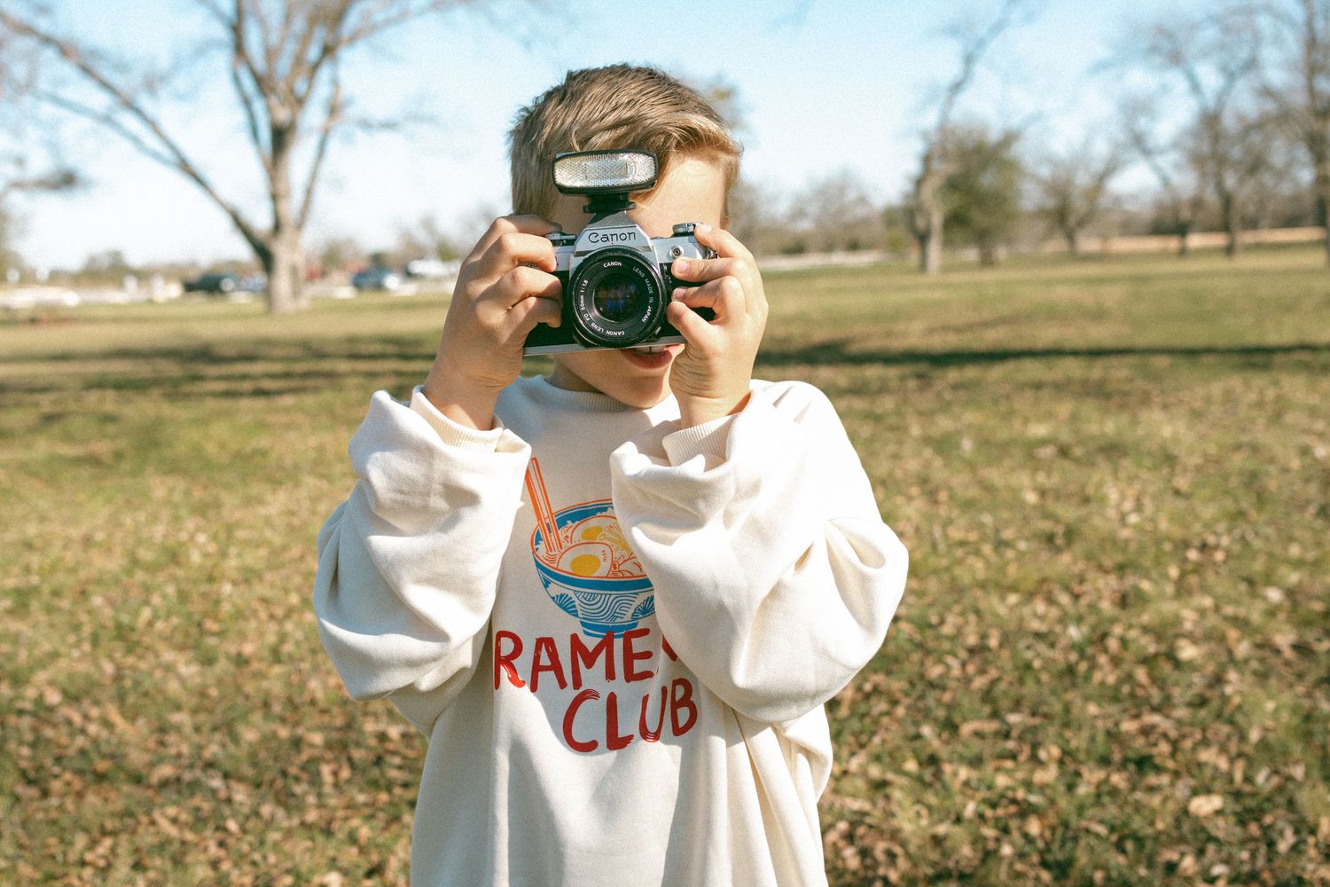 Person in a white sweater with RAME CLUB text holds a vintage camera while standing in a sunlit autumn field.