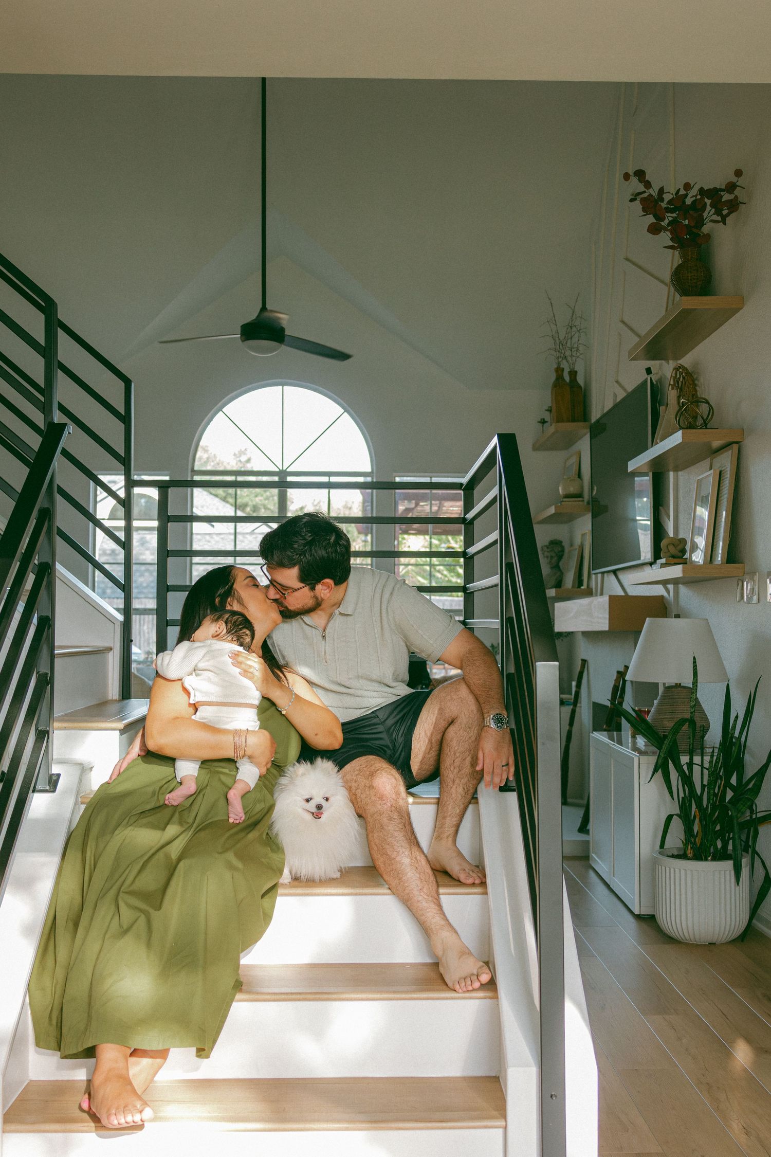 Casual in-home lifestyle newborn session with parents kissing on stairs covered in dappled light with a small, white Pomeranian.