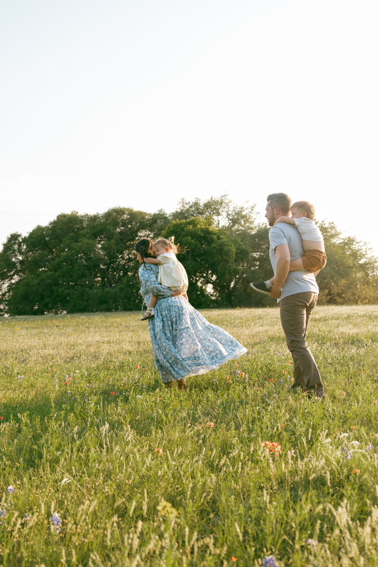 A couple carrying their two children in piggyback rides in a sunlit meadow during golden hour.