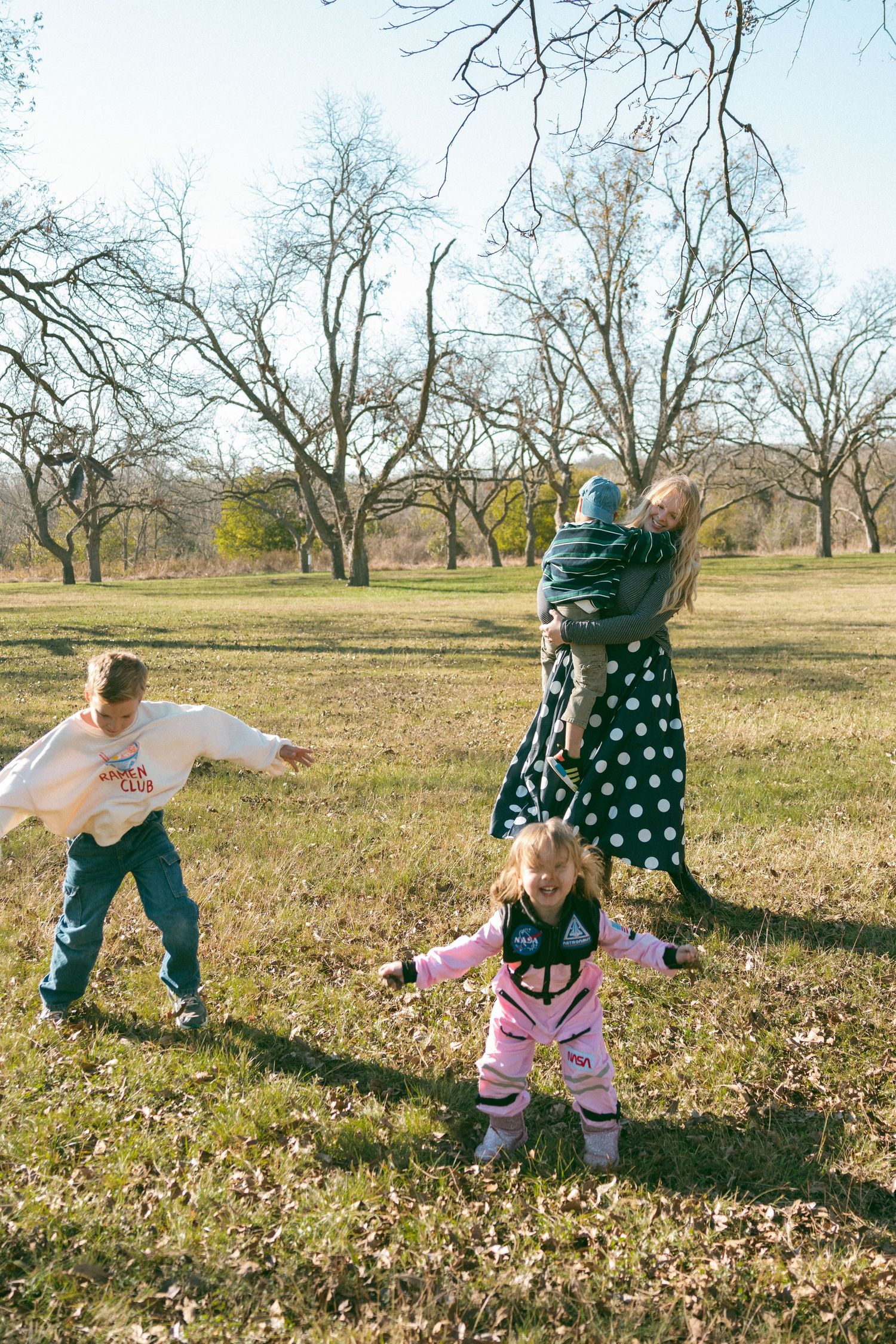 Children playing and rolling around in fallen autumn leaves on a sunny day with bare trees in the background.
