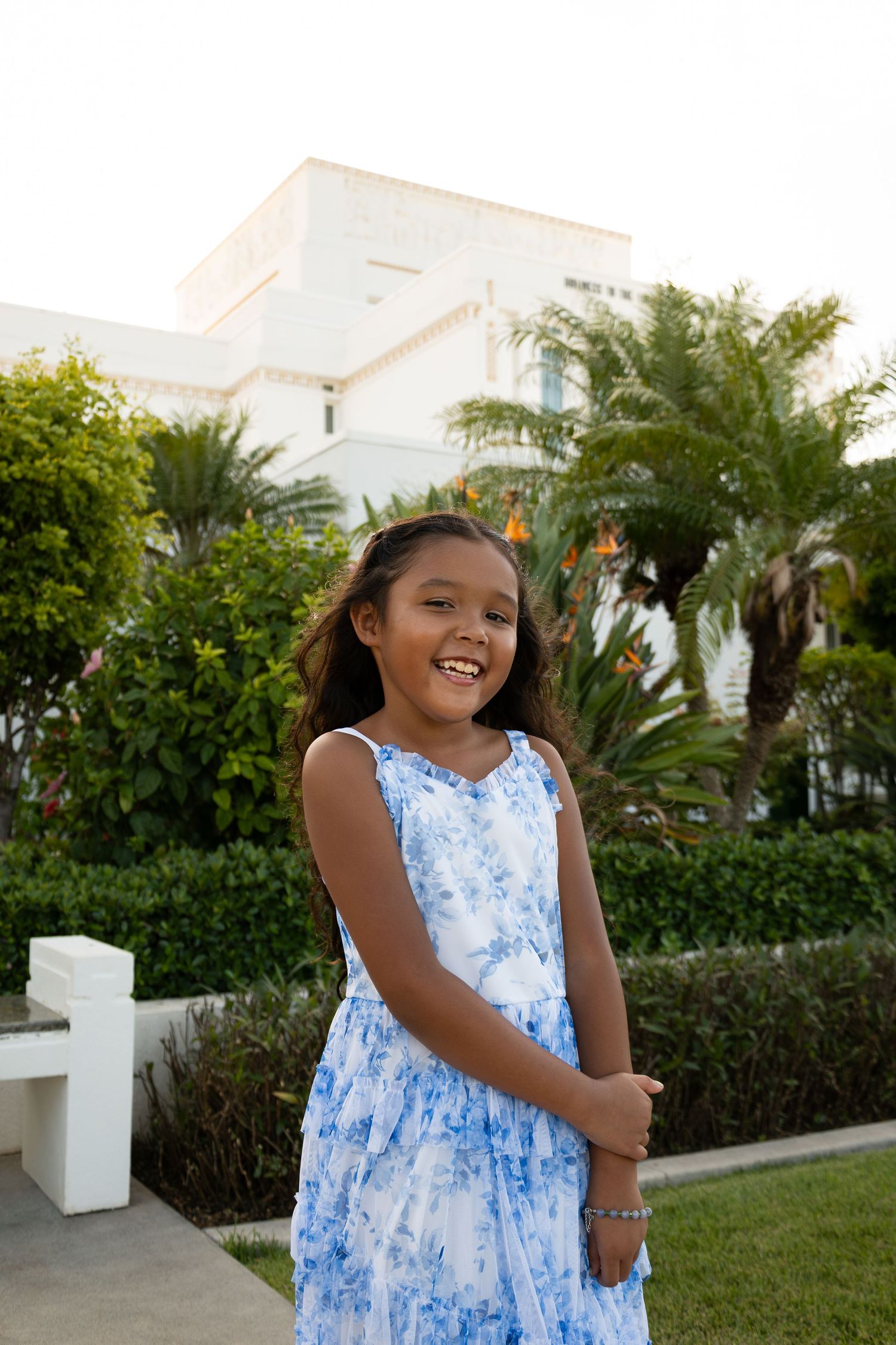 Smiling young girl in blue floral dress posing outdoors near tropical garden and white building.