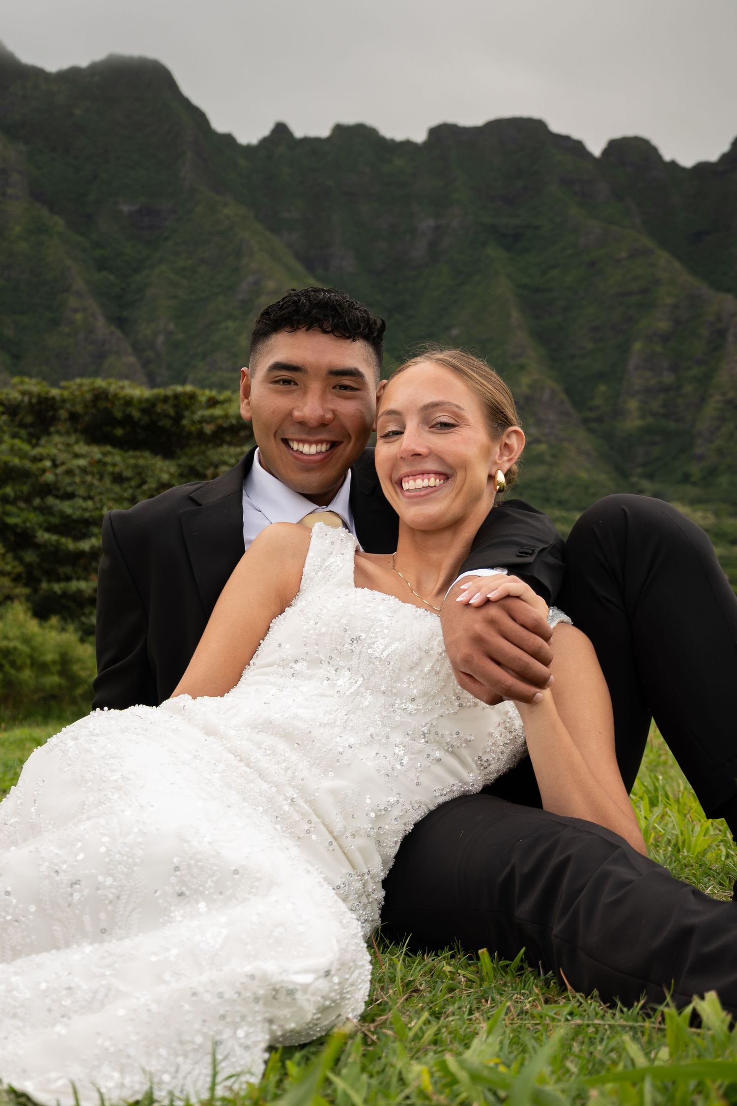 Bride and groom sitting on grass laughing, with lush green mountains backdrop, Hawaii wedding portrait.