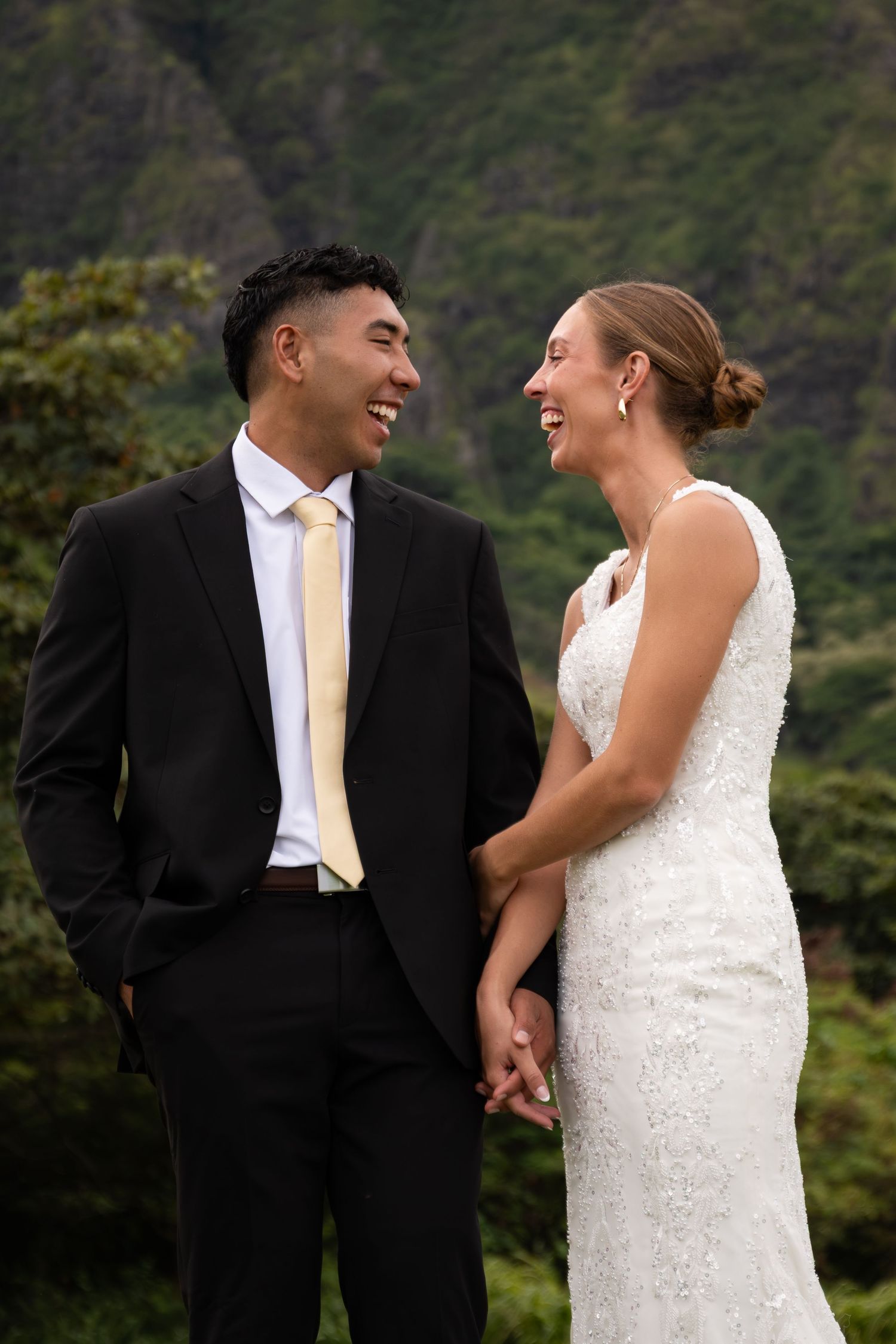 A laughing bride and groom hold hands outdoors with lush green mountains behind them on their wedding day.